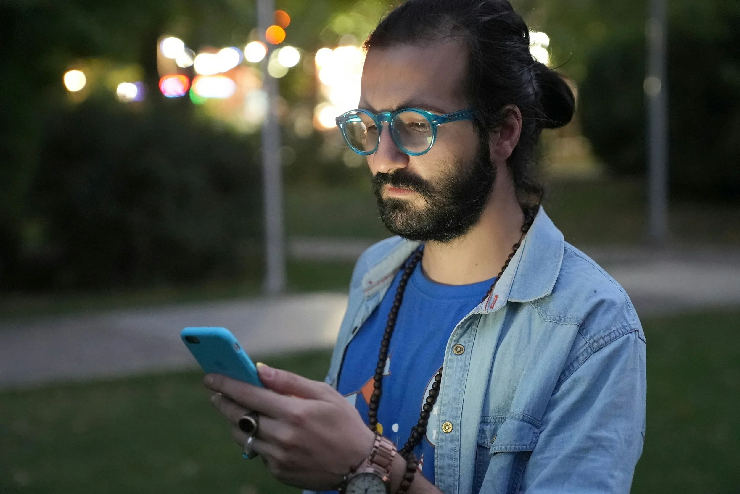 A bearded man with glasses and a hair bun looks at his phone outdoors in the evening.
