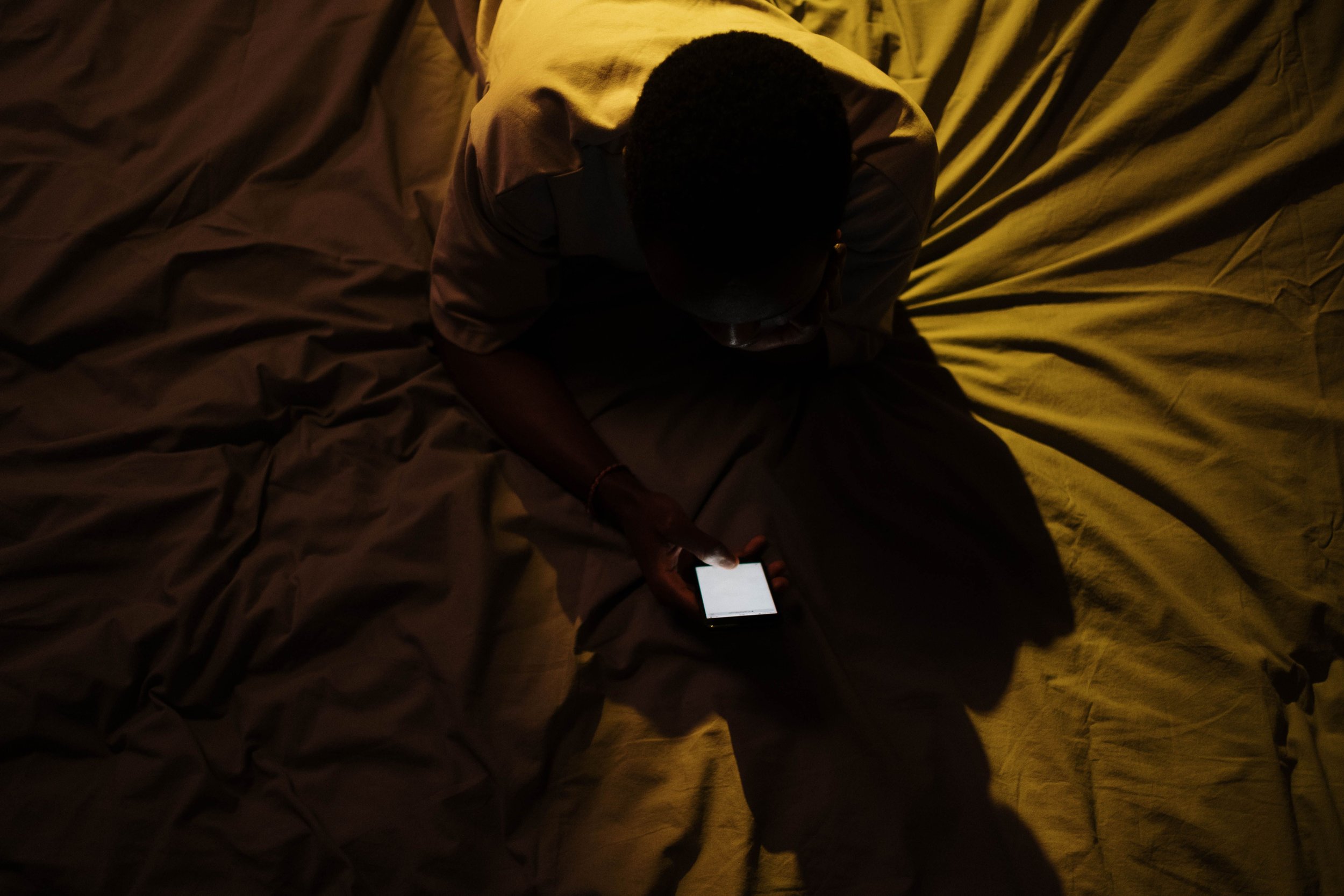 A person sitting on the floor surrounded by crumpled blankets or sheets, looking at a smartphone in their hand in a dimly lit room.