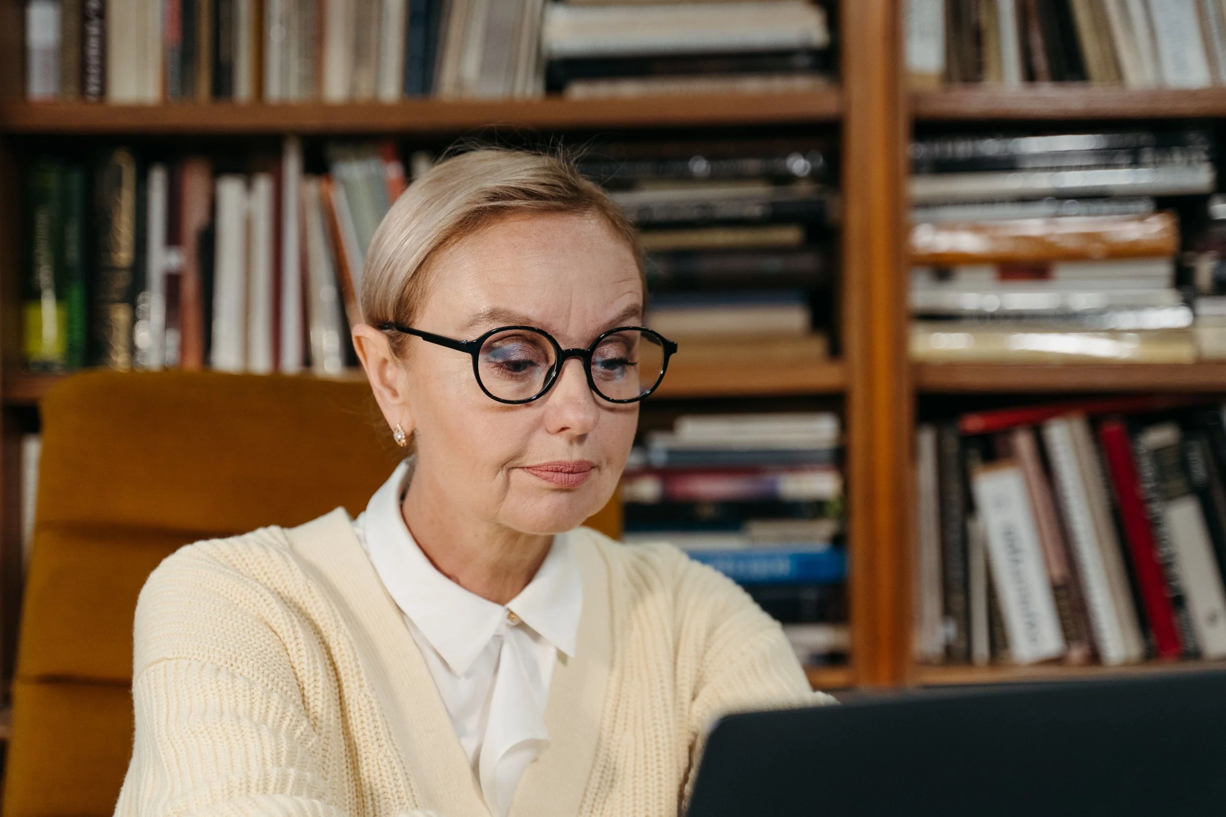 An older woman with glasses and blonde hair looks at a laptop, seated in front of a large bookcase.