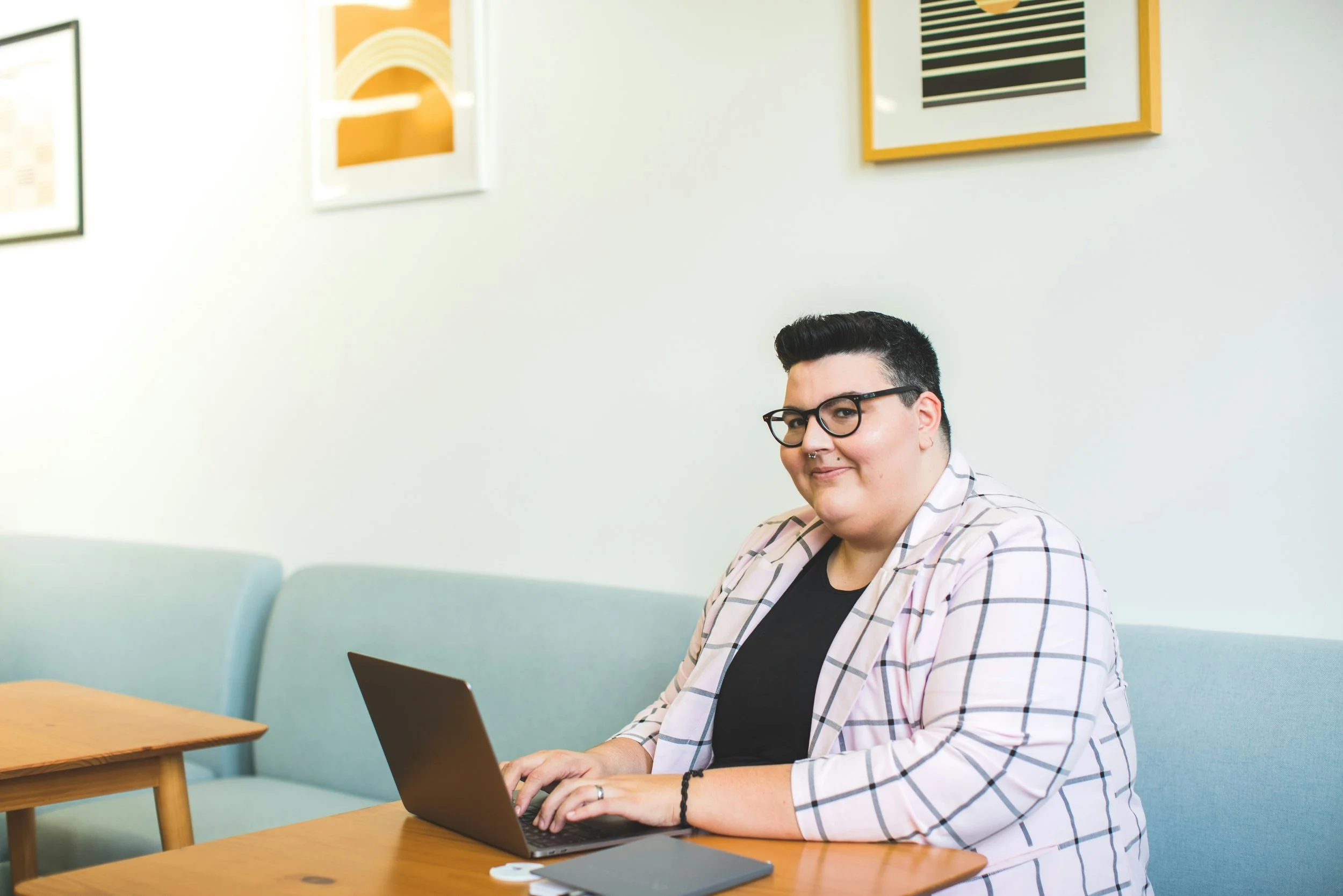 A person with short dark hair and glasses smiles at the camera while working on a laptop in a café.