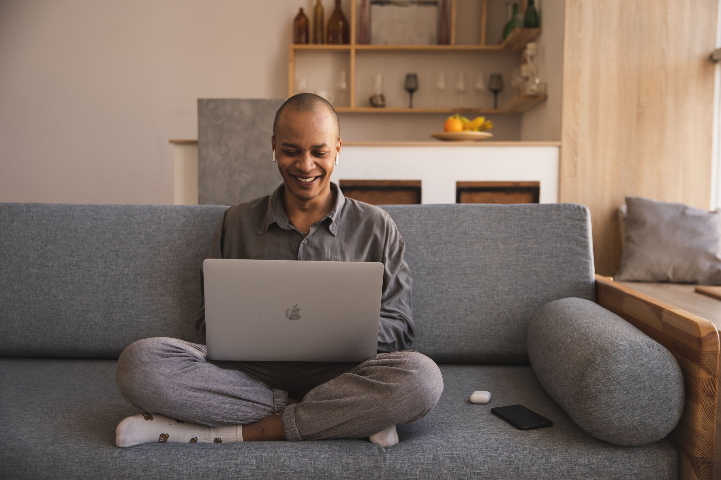 A smiling man sits cross-legged on a grey sofa, using a laptop with headphones in.