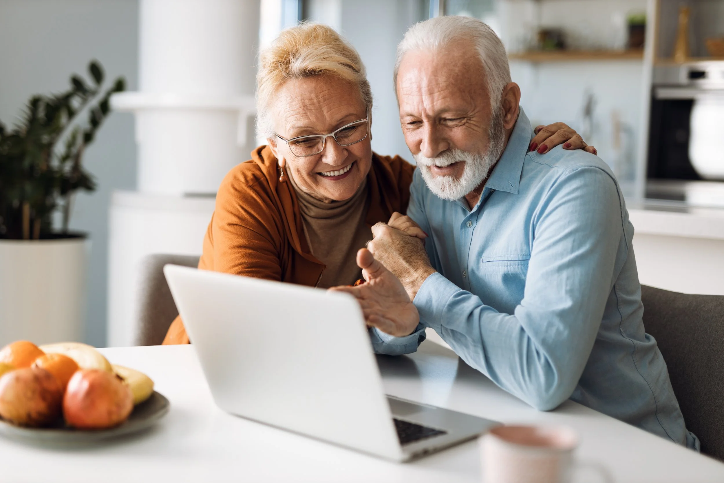 Senior couple smiling and embracing while looking at a laptop screen in a bright kitchen.