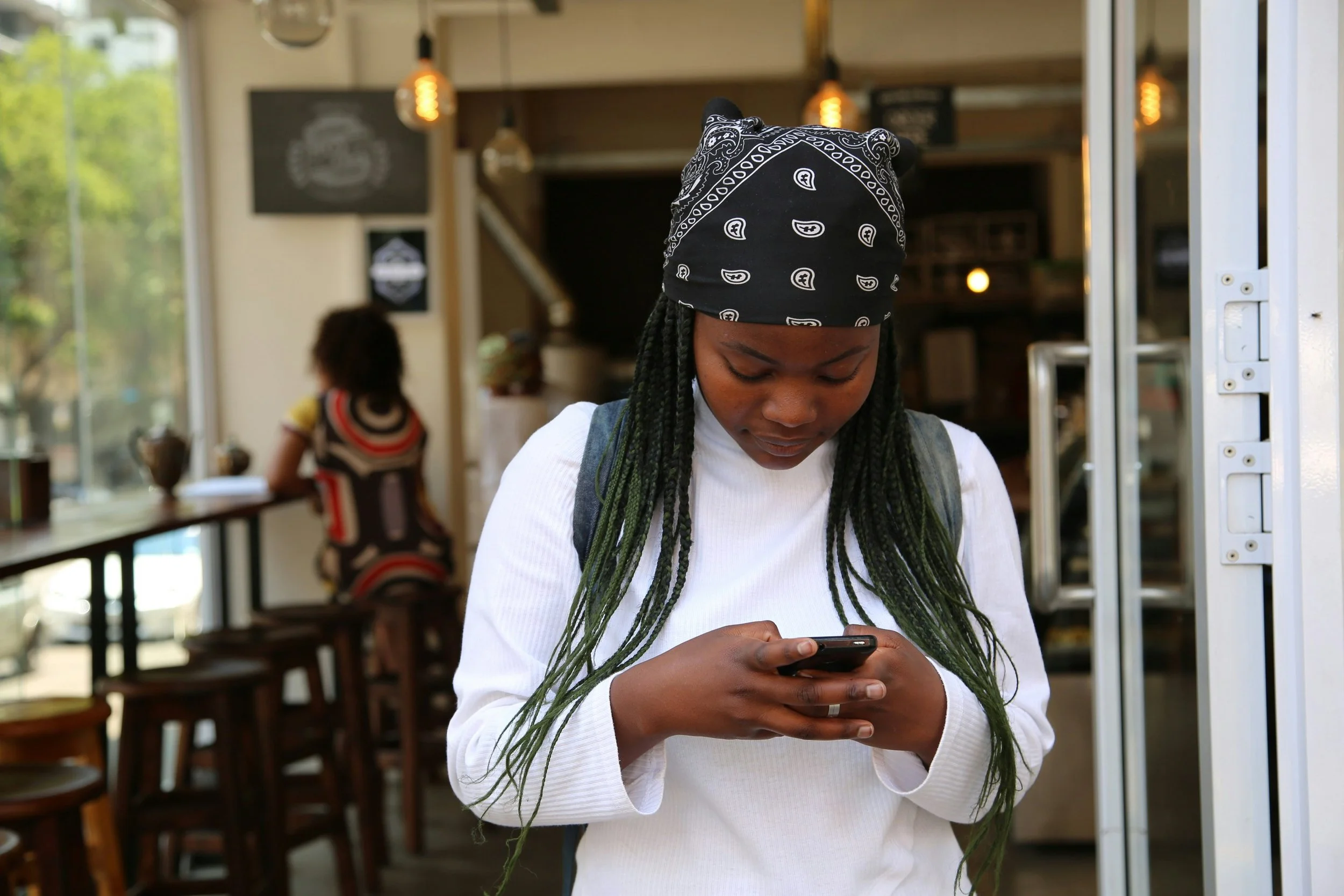 A woman with long braids and a bandana looks down at her phone while standing in a café doorway.