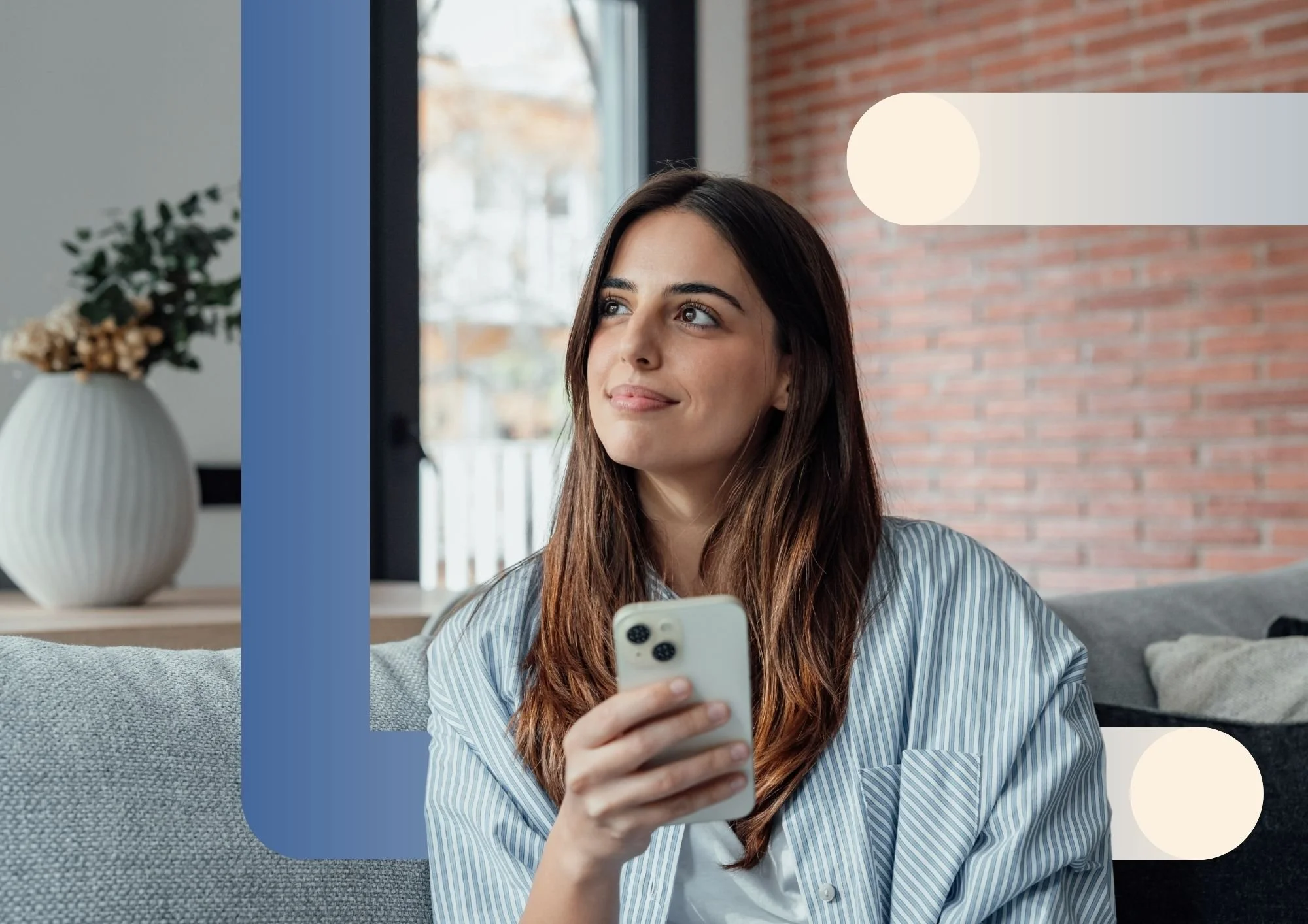A woman sitting on a gray sofa holding a smartphone, with a brick wall and large window in the background, in a cozy living room setting.
