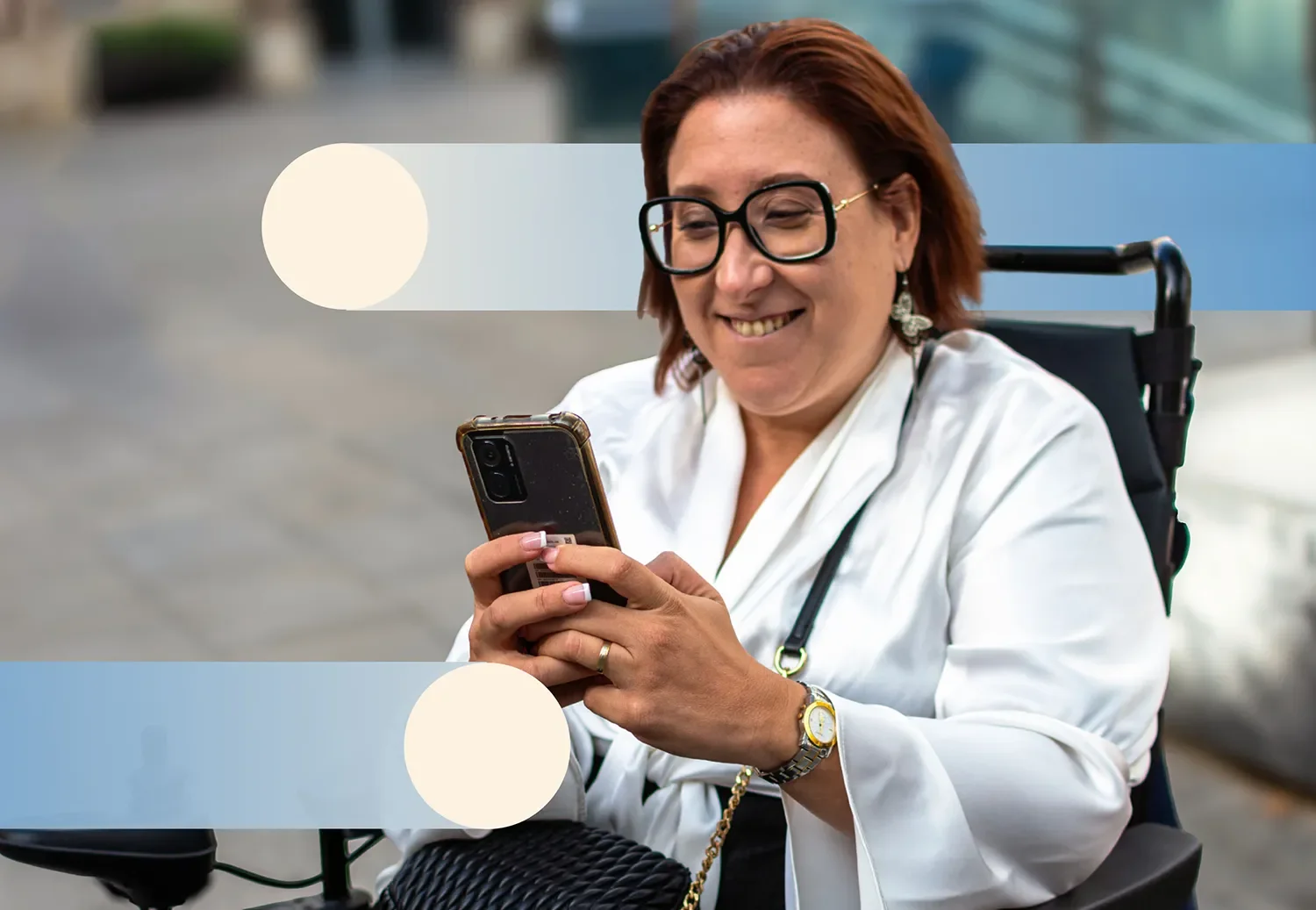 A woman in a wheelchair, smiling and looking at her phone, with short red hair, wearing glasses, a white jacket, and earrings, outdoors in an urban setting.