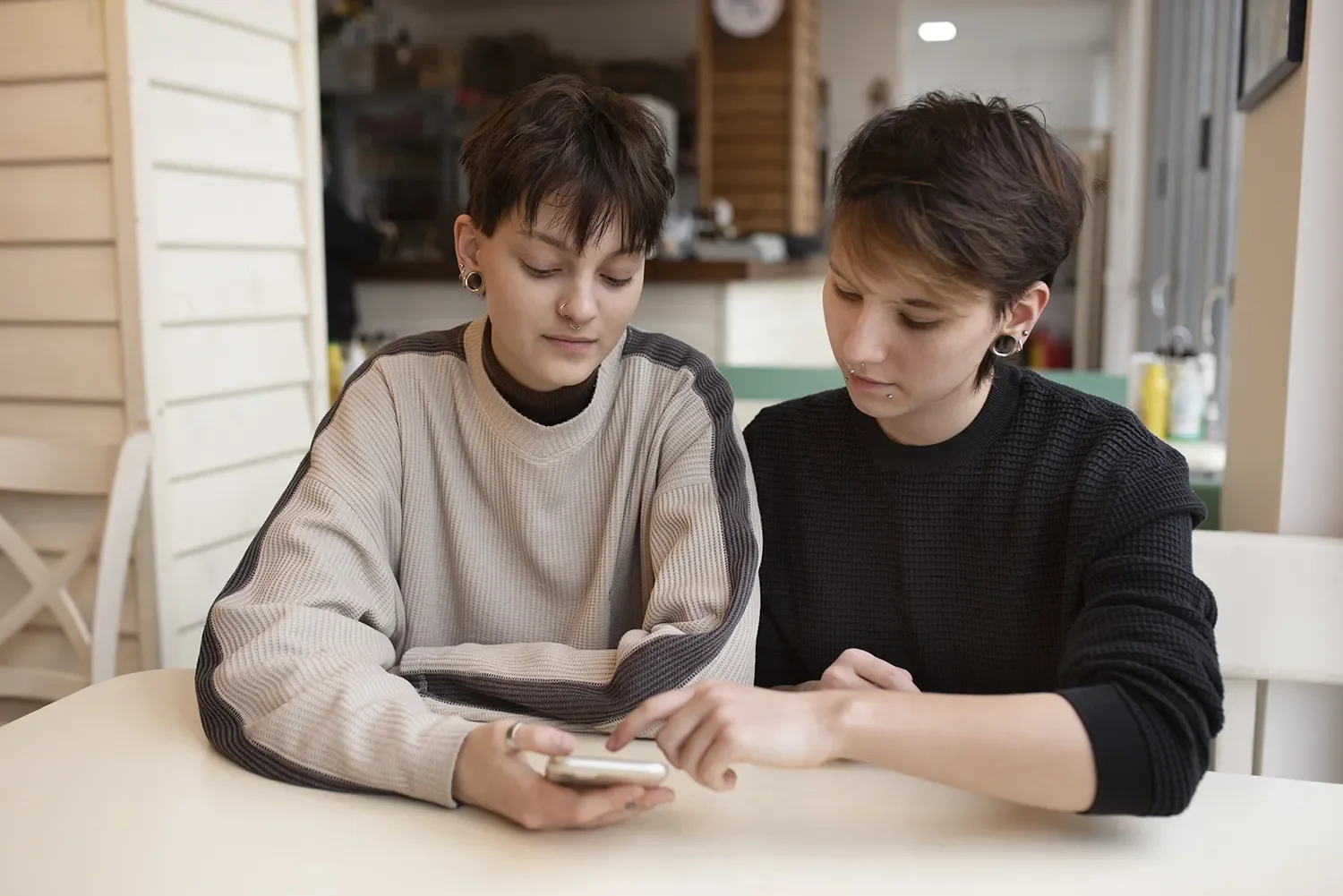 Two young people with short dark hair and piercings sitting at a white table, looking at a smartphone together in a cozy kitchen or cafe setting.