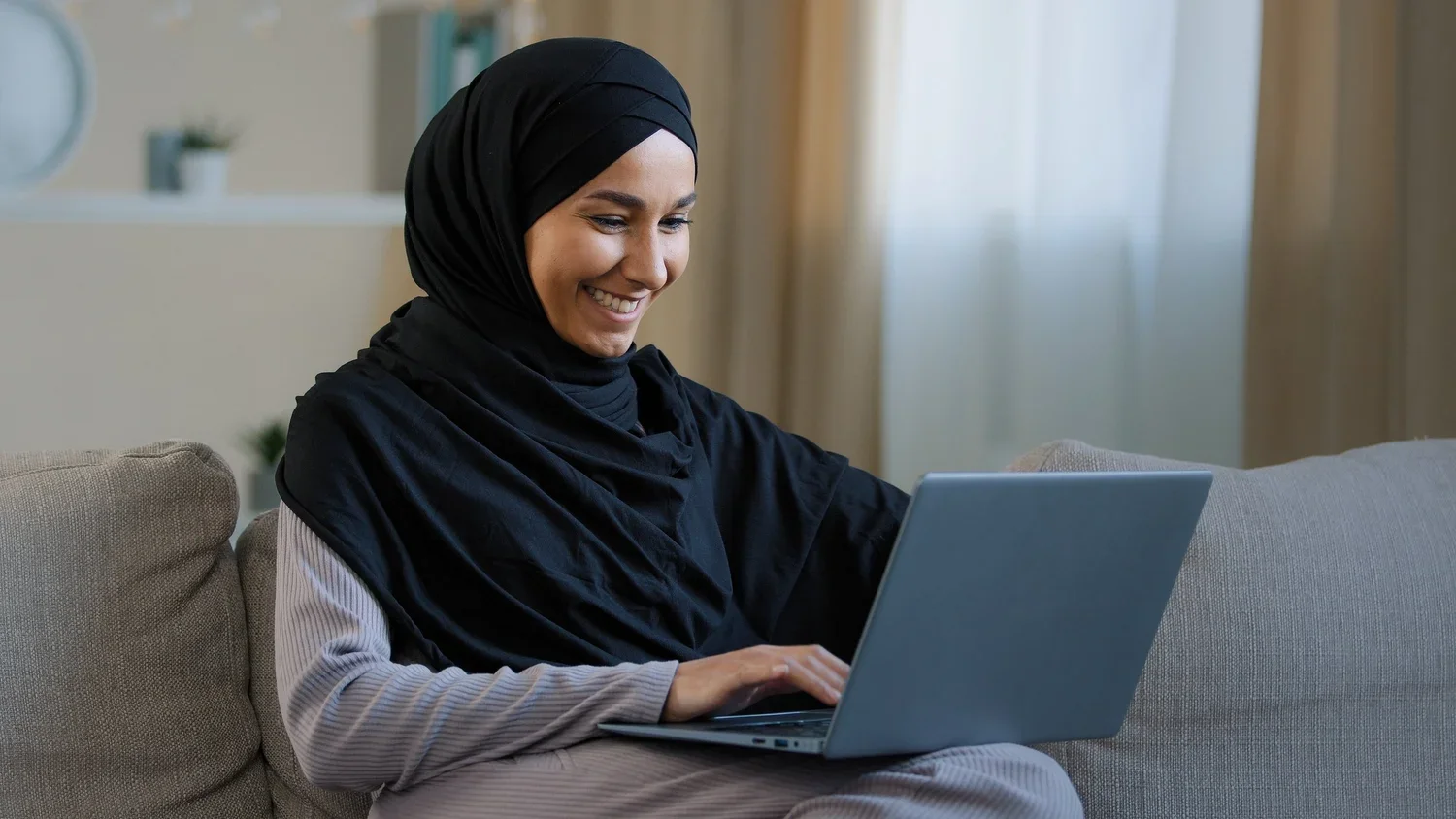 a woman in a black hijab smiling while using a gray laptop on a couch