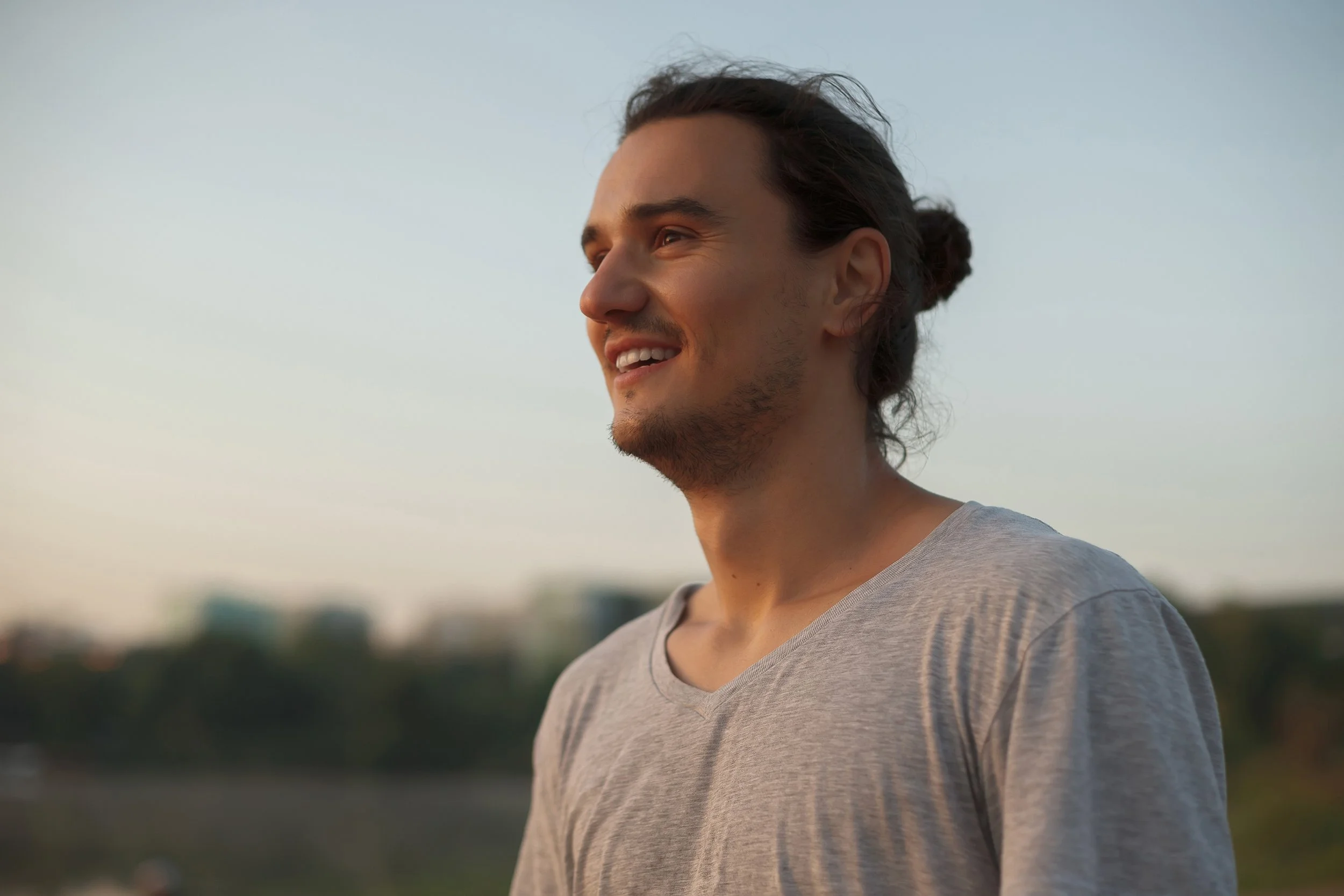 A young man with long, dark hair tied in a bun, smiling and looking off into the distance during the late afternoon or early evening outdoors.
