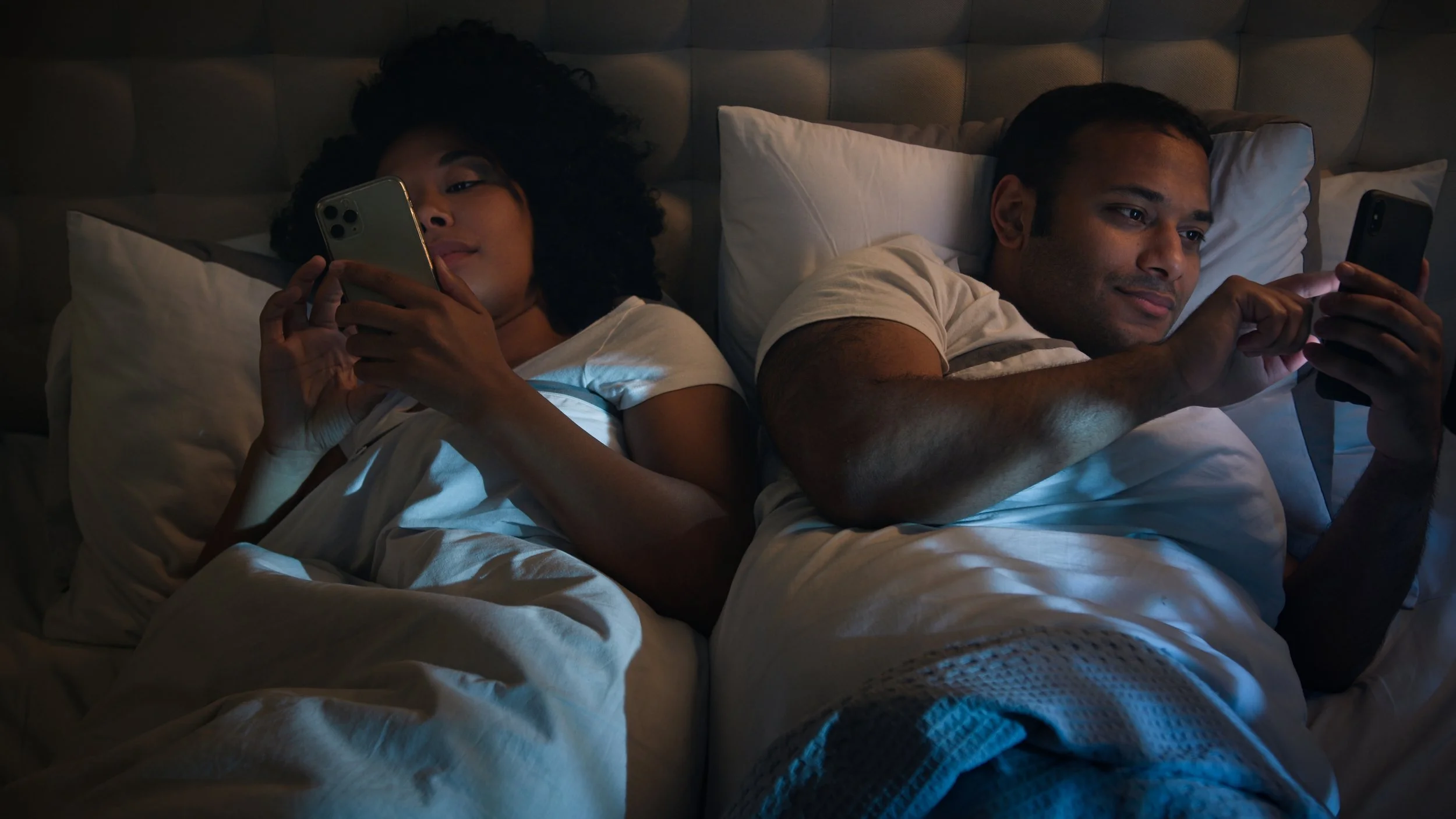 Two people lie in bed at night, each looking at their own phone screen in the dark.