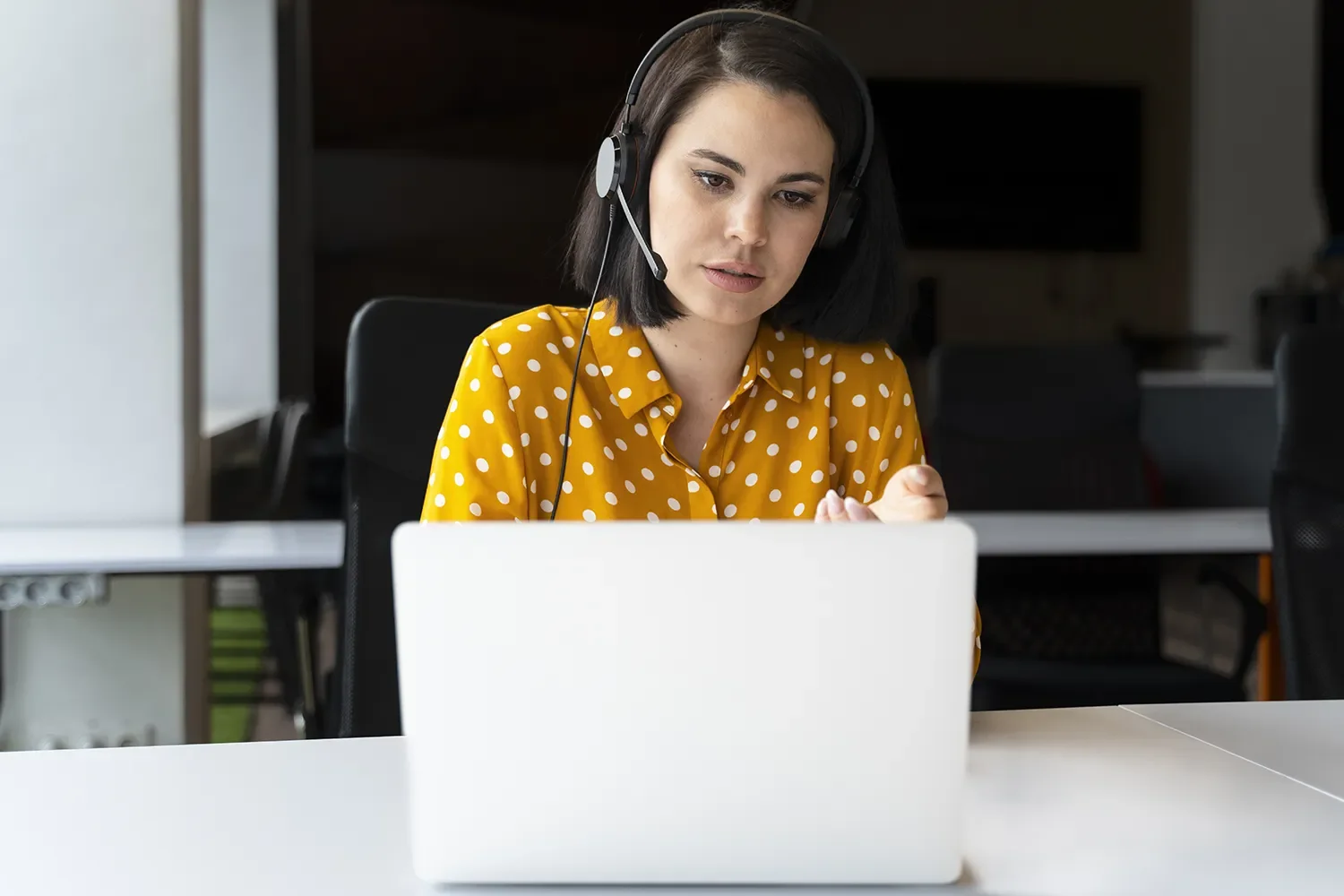 A woman with dark hair wearing a yellow polka dot shirt using a headset while working on a laptop in an office.