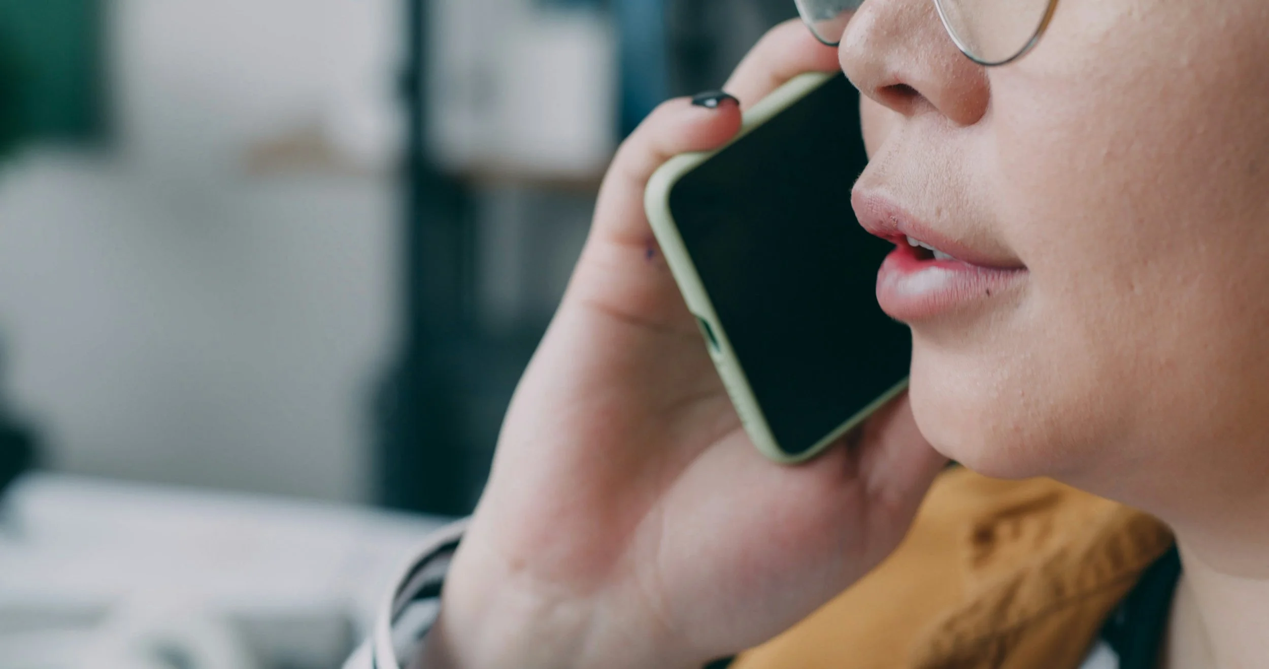 A close-up of a person holding a phone to their mouth, wearing a mustard yellow top.