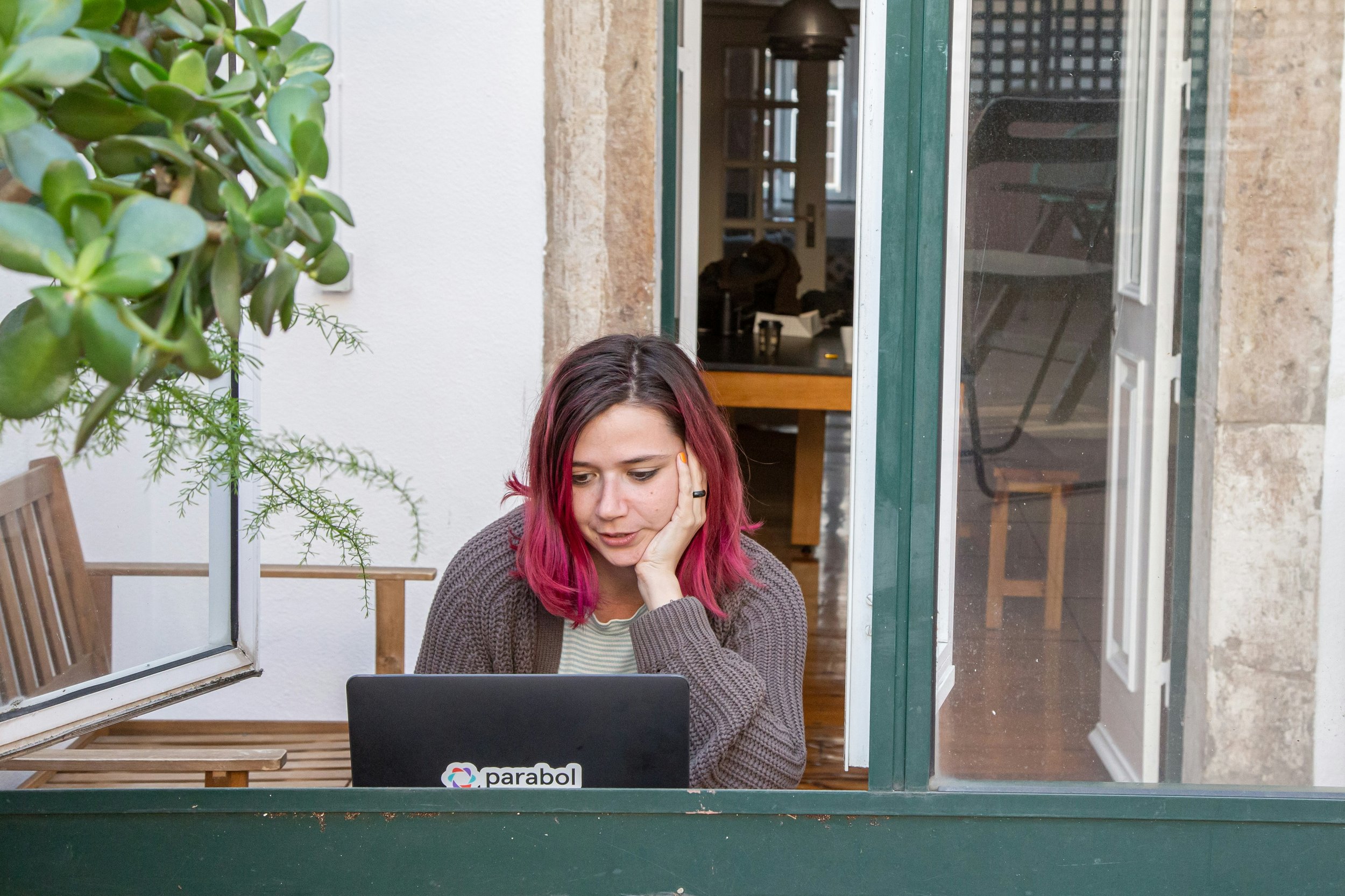 A woman with pink ombre hair sits at a table, focused on her laptop, with a cup of coffee nearby.