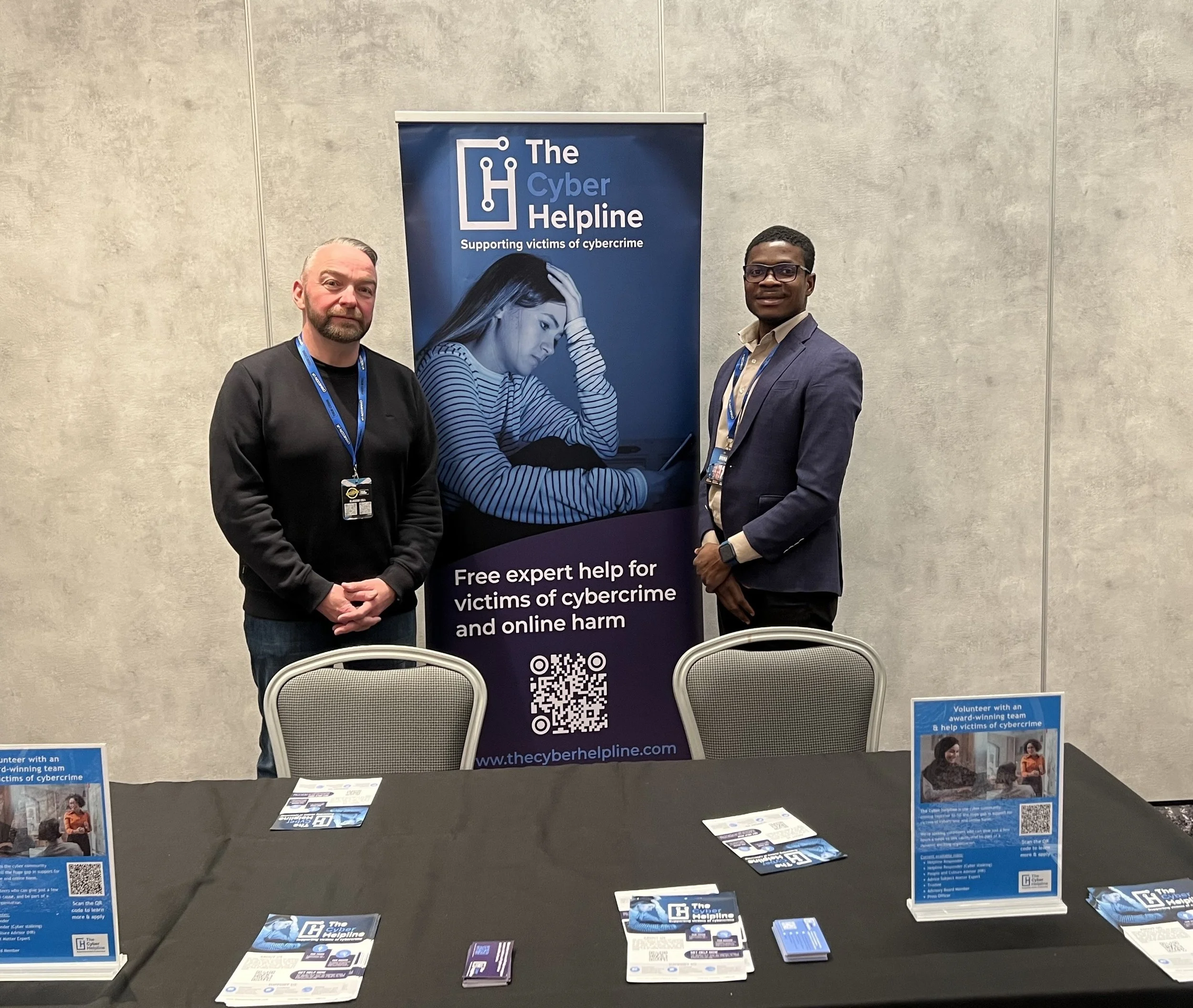 Two men standing next to a blue banner that reads 'The Cyber Helpline, Supporting victims of cybercrime.' The banner features an image of a woman with her hand on her forehead and a QR code at the bottom. The men are wearing conference badges and are positioned in a room with a plain wall in the background.