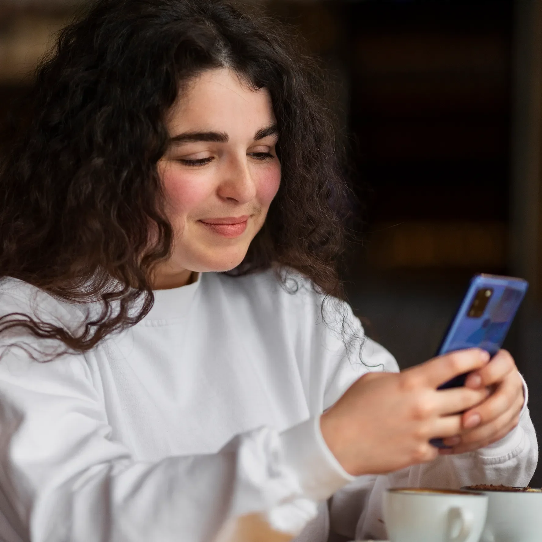 A young woman with curly dark hair smiling and looking down at her phone, which she is holding with both hands. She is wearing a white shirt.