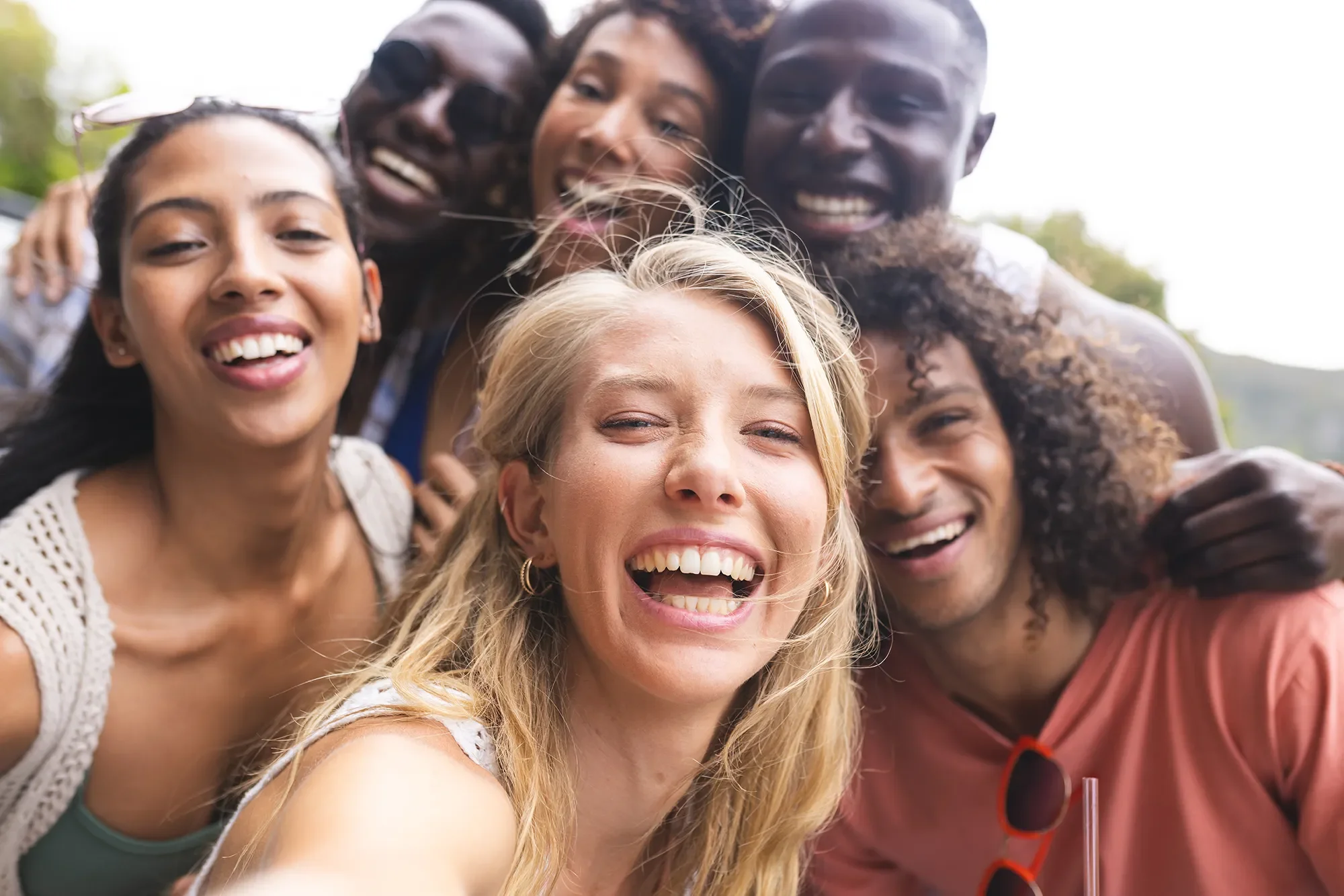 A group of six diverse friends smiling and taking a selfie together outdoors.