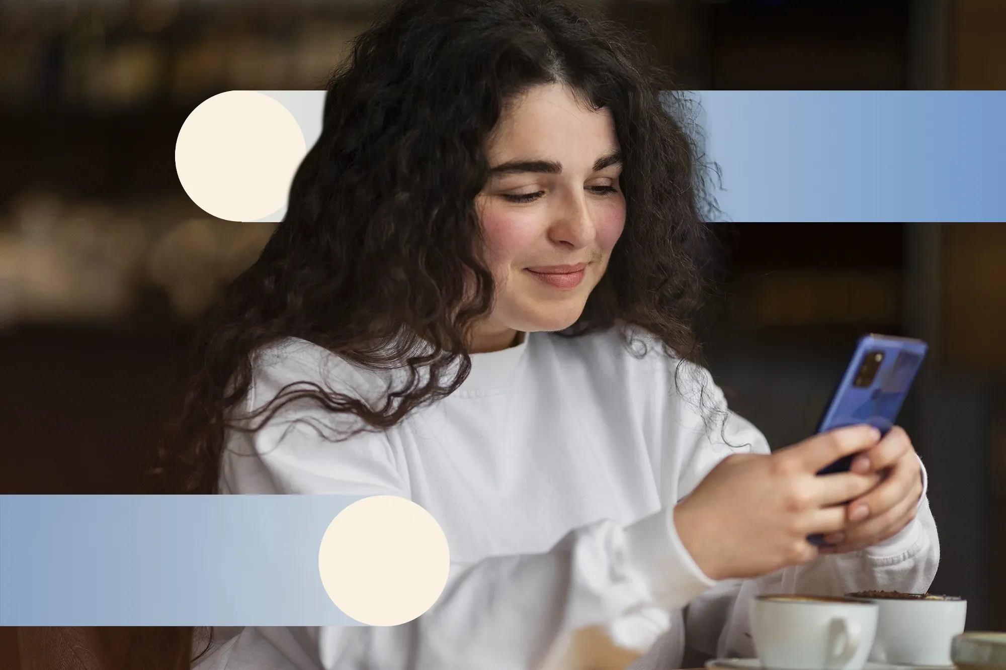 A young woman with curly brown hair smiling while looking at her smartphone, sitting at a table with a cup in front of her.