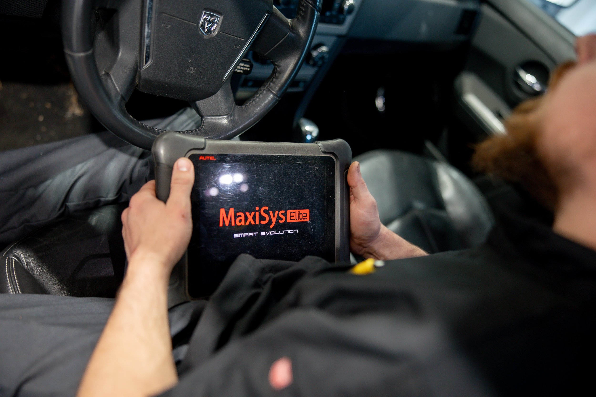 Person holding a diagnostic scan tool labeled 'MaxiSys Elite' inside a vehicle, with the steering wheel and dashboard visible.
