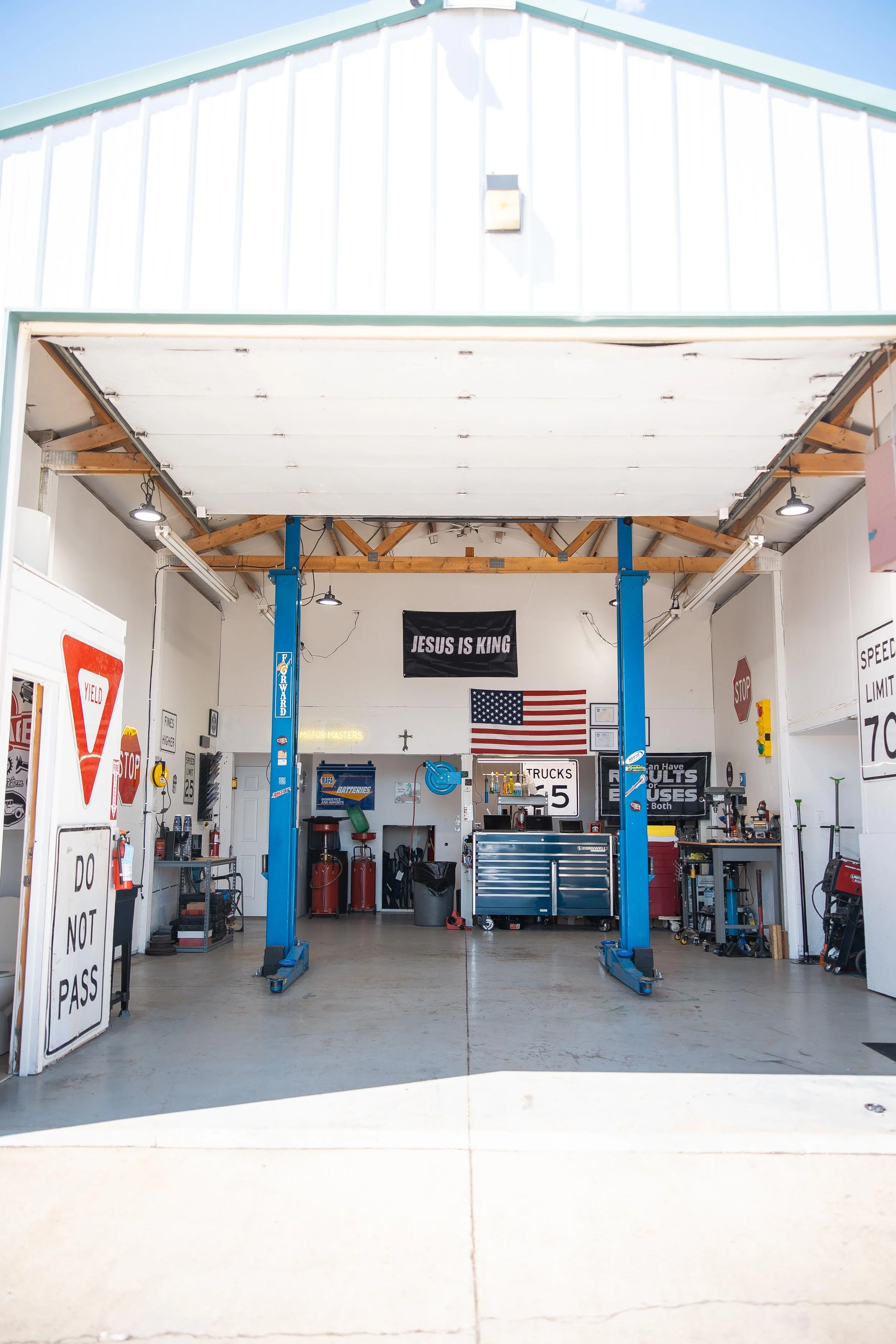 The interior of Idaho Motor Masters, an auto repair garage with a vehicle lift, tools, and signs, including a black banner that reads 'JESUS IS KING', an American flag, and various garage equipment.