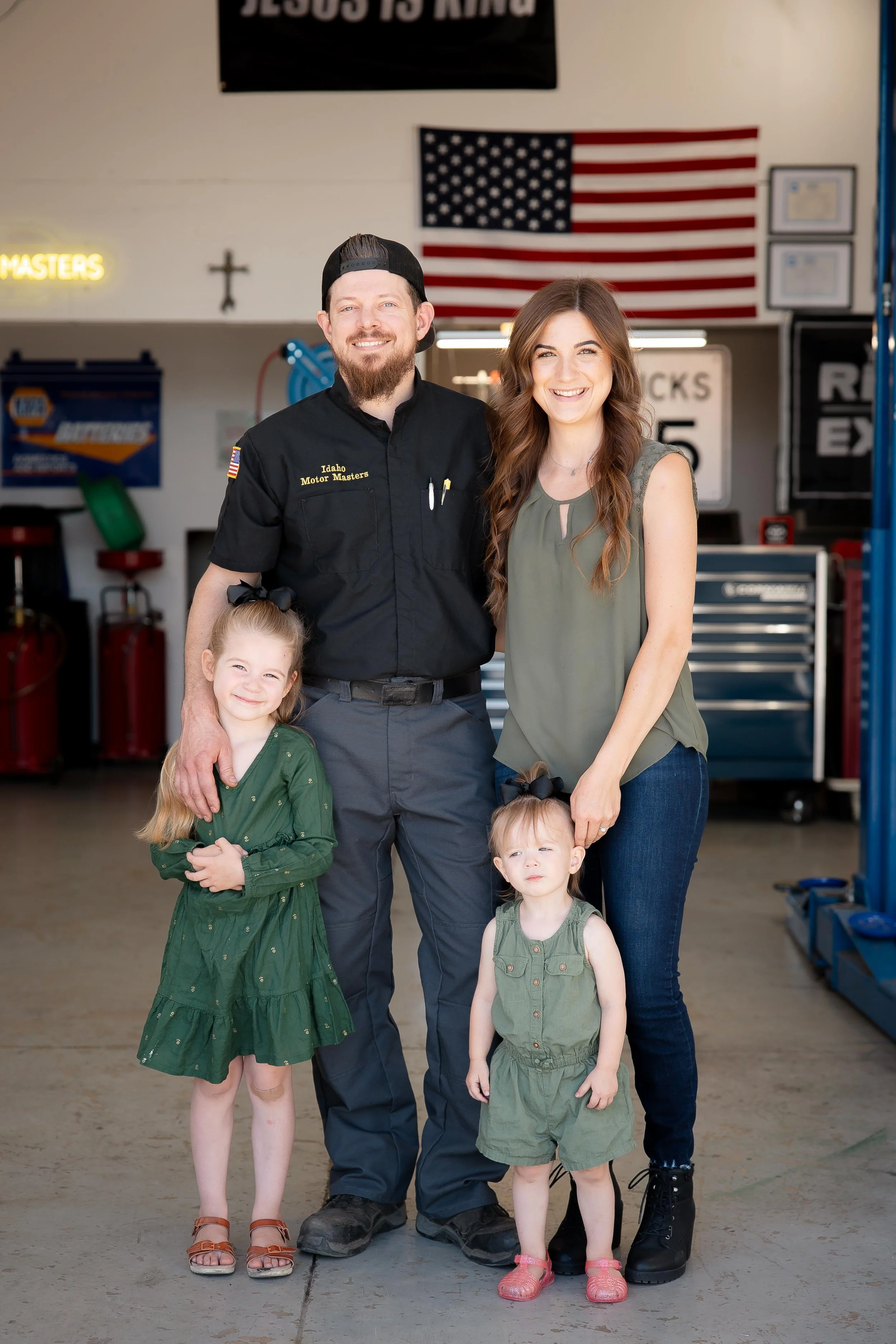 Eldon Johnson, Idaho Motor Master Shop Owner & his wife and two daughters. A family oriented man with honest care. A family of four smiling and posing in a garage or workshop with American flags and automotive signs in the background.