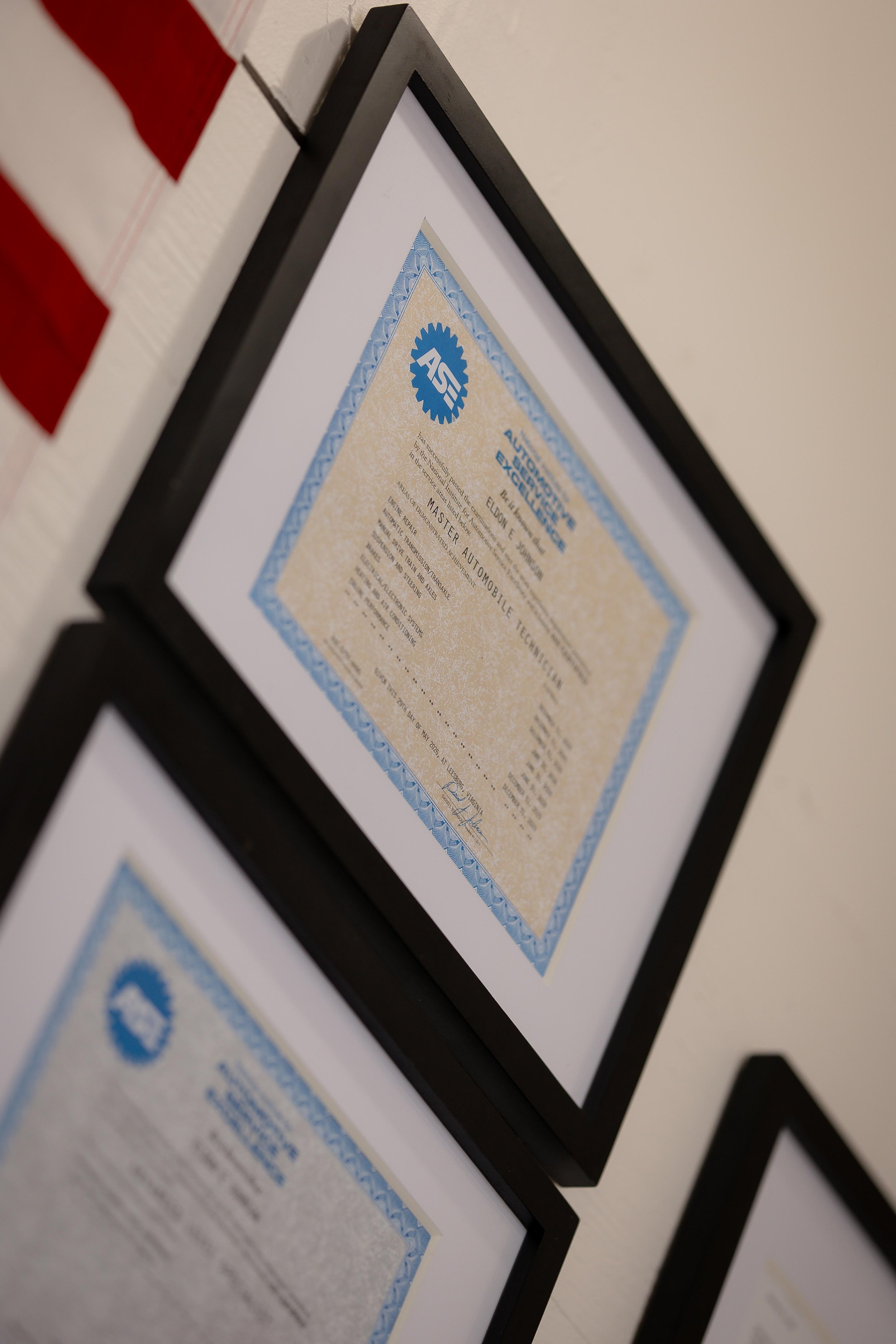 Framed certificate with a blue border hanging on a wall. The certificate is issued by the ASE (Automotive Service Excellence) and is for a Master Automobile Technician.