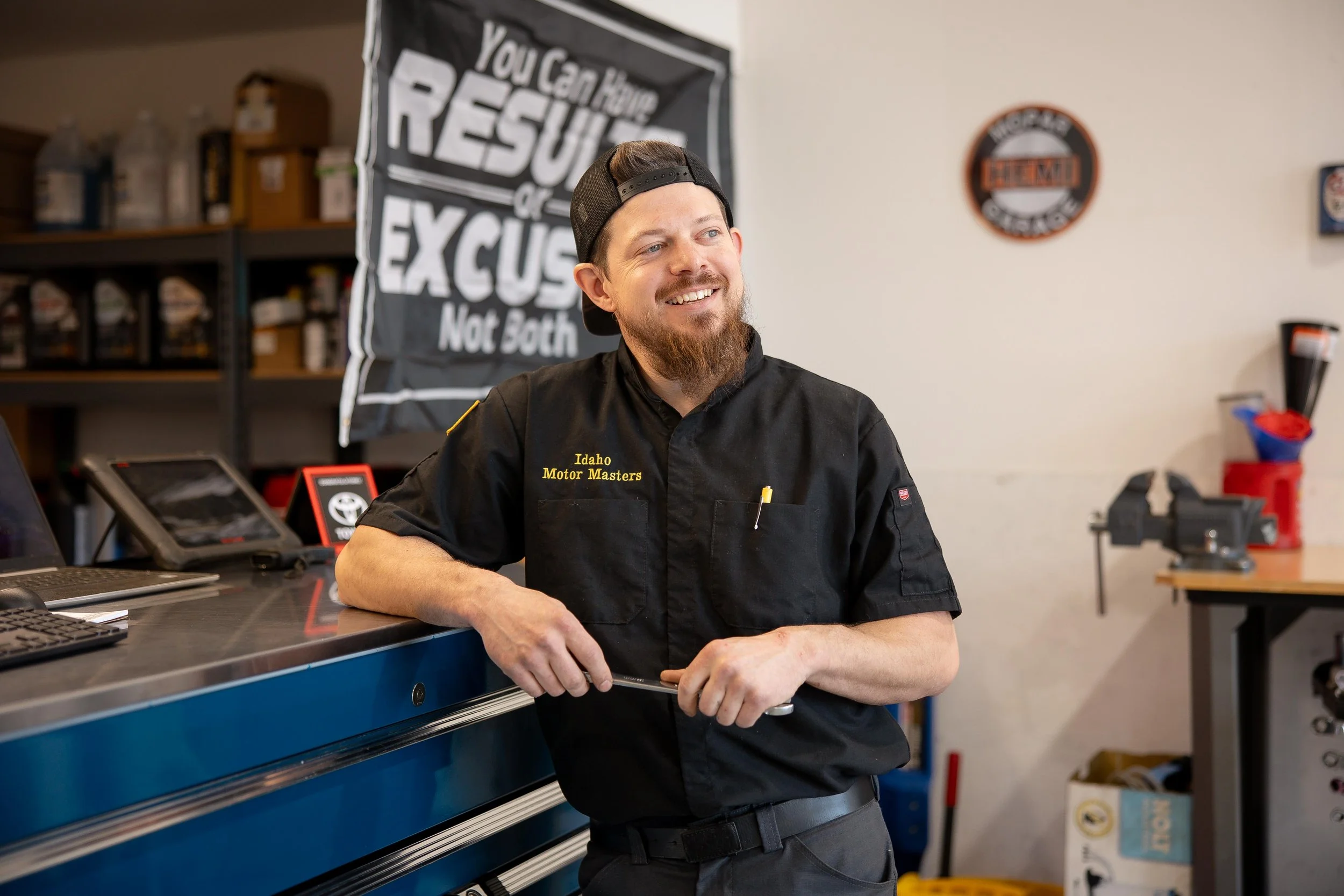 A man with a beard and a backwards baseball cap smiling and leaning on a workbench in a workshop. He's wearing a black shirt with yellow lettering that says 'Idaho Motor Masters.' In the background, there is a black cloth banner with white text reading 'RESUME or EXCUSE Not Both,' some shelves with bottles and boxes, and a white wall with a round sign.
