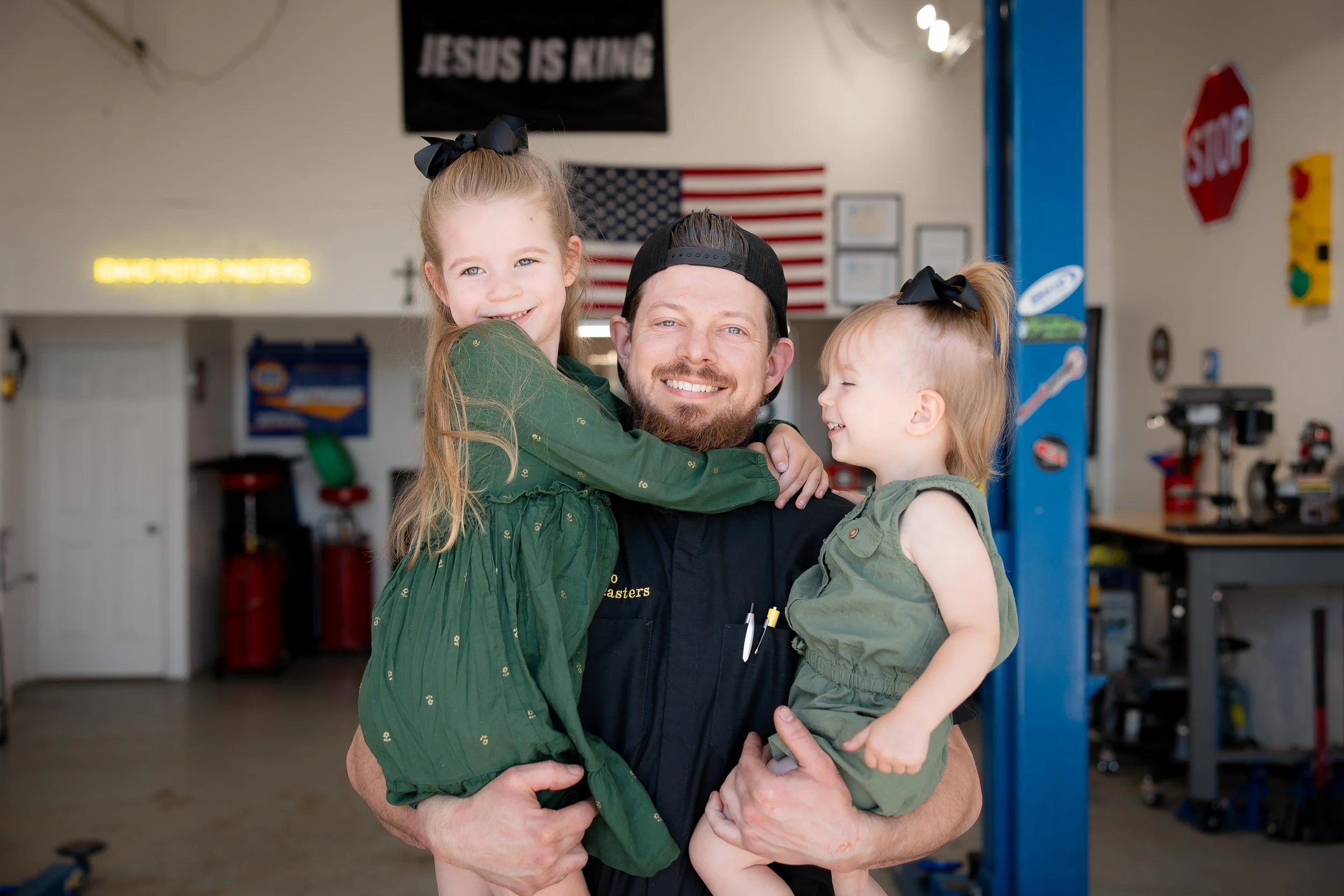 A man holding two young girls, one on each arm, inside a garage or workshop with American flags and safety signs in the background.
