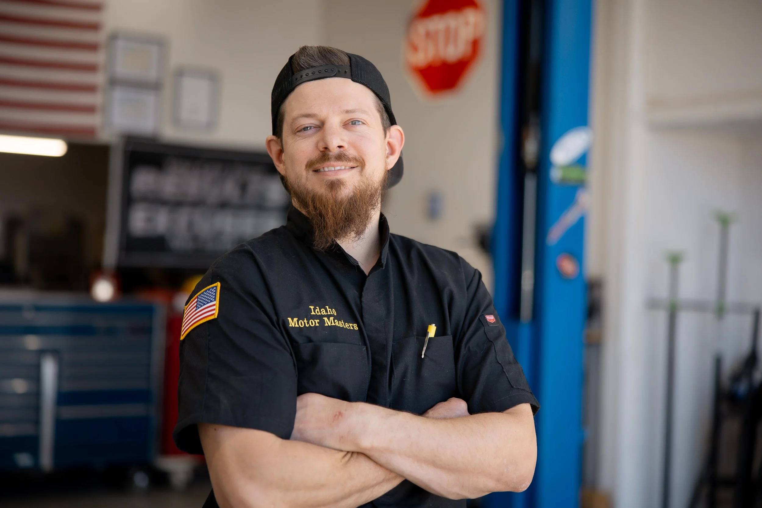 A smiling man with a beard wearing a black cap backward and a black uniform with an Idaho Motor Masters patch, standing with arms crossed in an automotive repair shop.