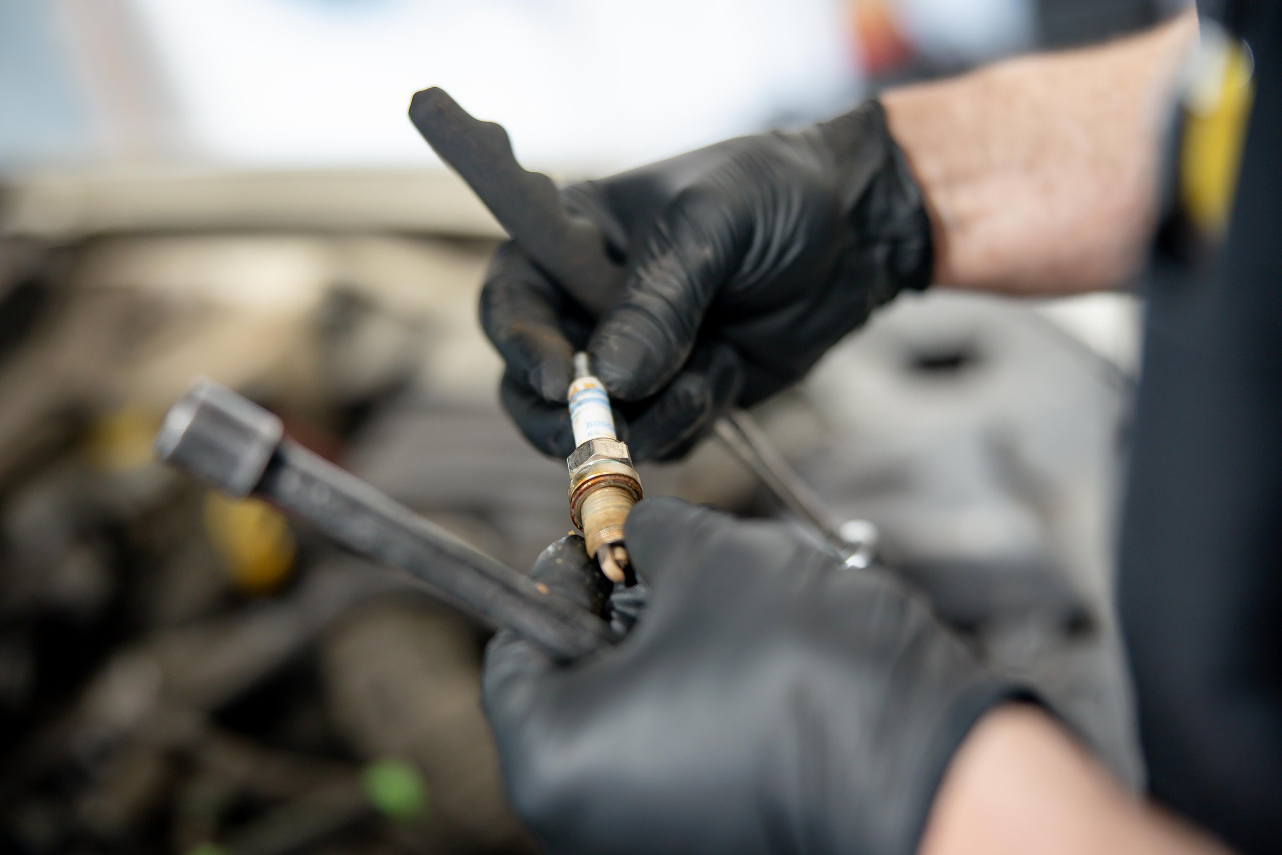 A person wearing black gloves holding a tool, working on a metallic component in a workshop or factory setting.