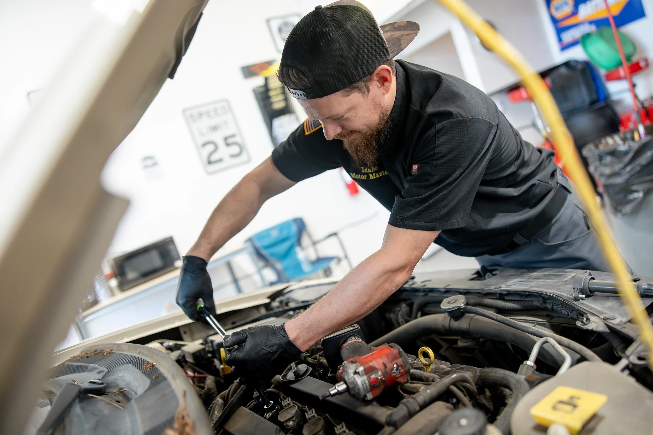 A man with a beard wearing a black cap backwards and a black mechanic uniform working under the hood of a car in a garage. There are tools and car parts visible around him.