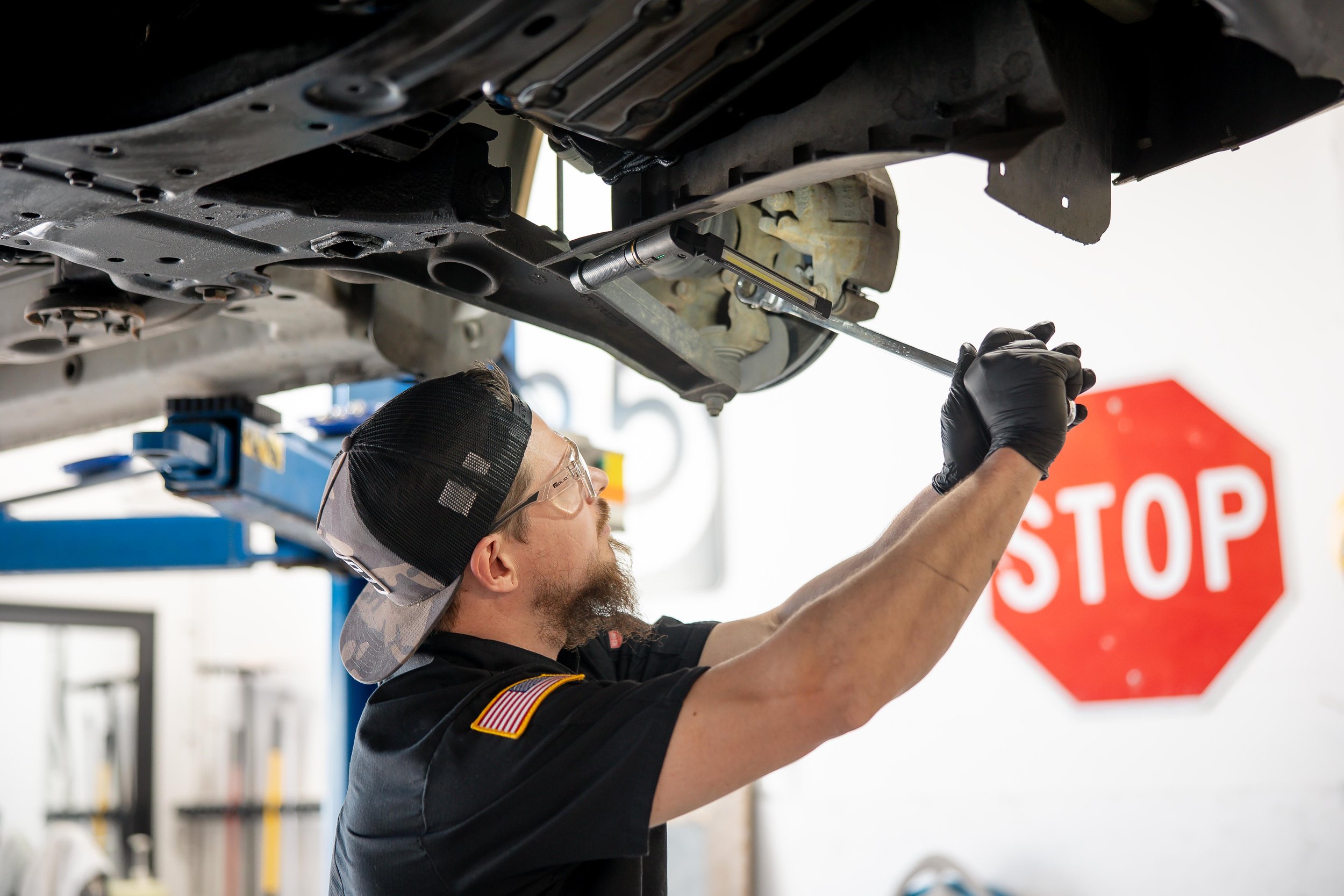 Eldon Johnson, lead mechanic wearing safety glasses, a cap, and black gloves is working underneath a vehicle, using a tool on the underside of the car in an auto repair shop.