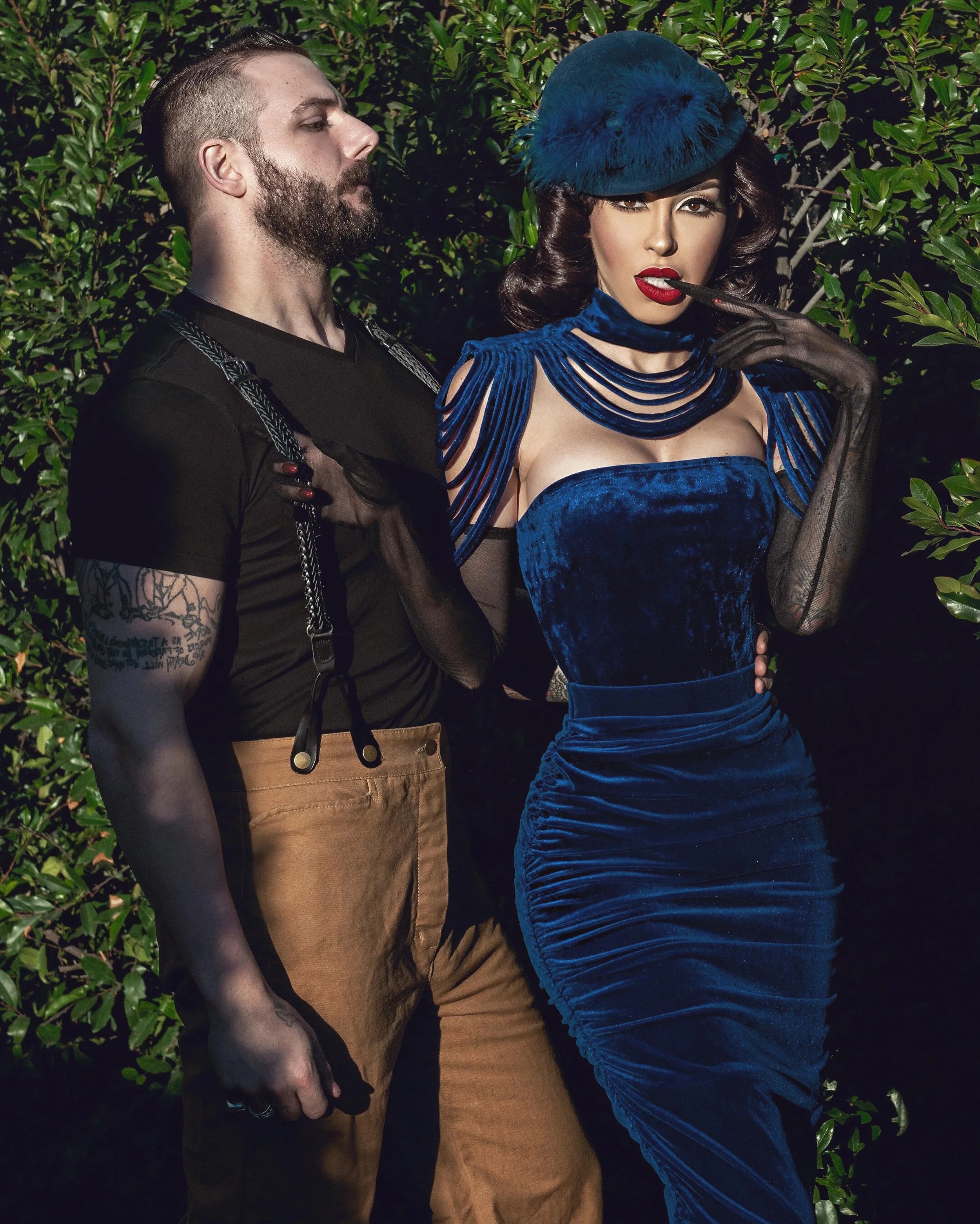 A man with a beard and tattoos on his arm stands next to a woman dressed in a royal blue velvet dress and matching hat, both posing outdoors in front of green bushes.