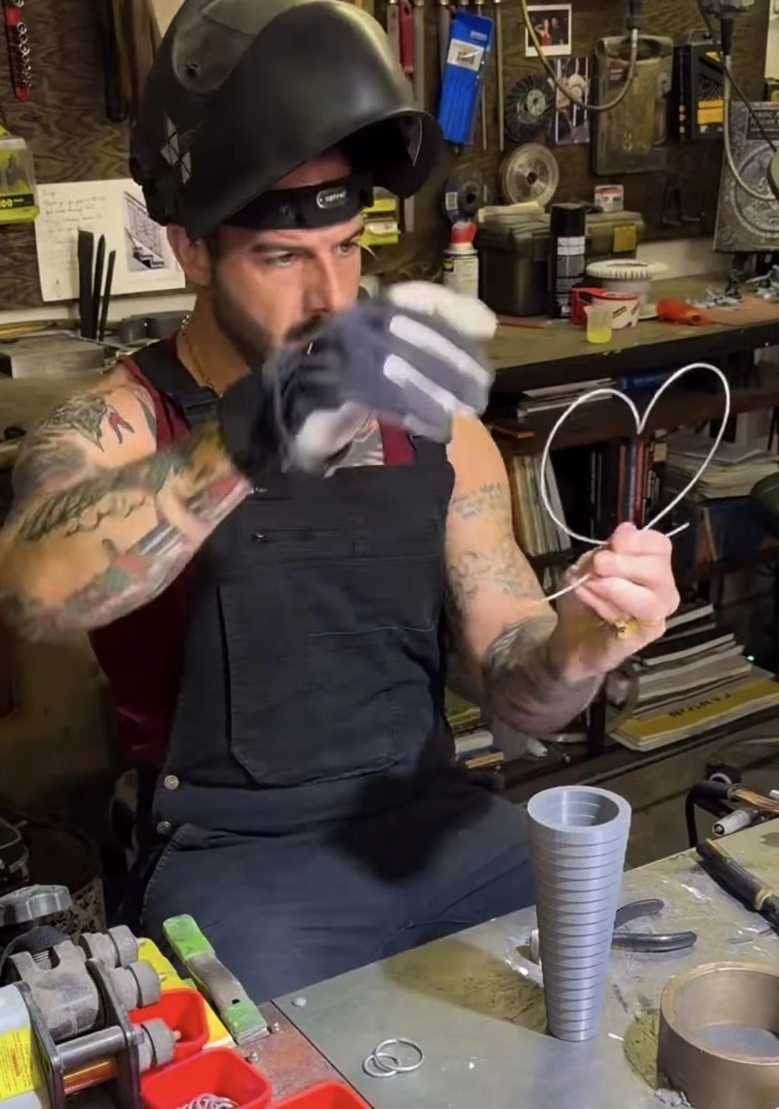 Man with tattoos wearing a motorcycle helmet and gloves, sitting at a workbench in a garage or workshop, shaping wire into a heart.