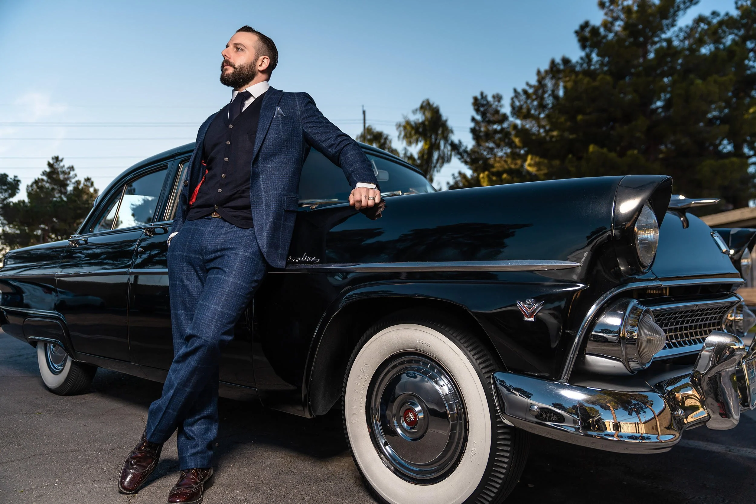 A man in a navy blue suit with a white shirt and dark tie, with a beard, standing beside a classic black Cadillac car with white-wall tires, outdoors with trees and power lines in the background.