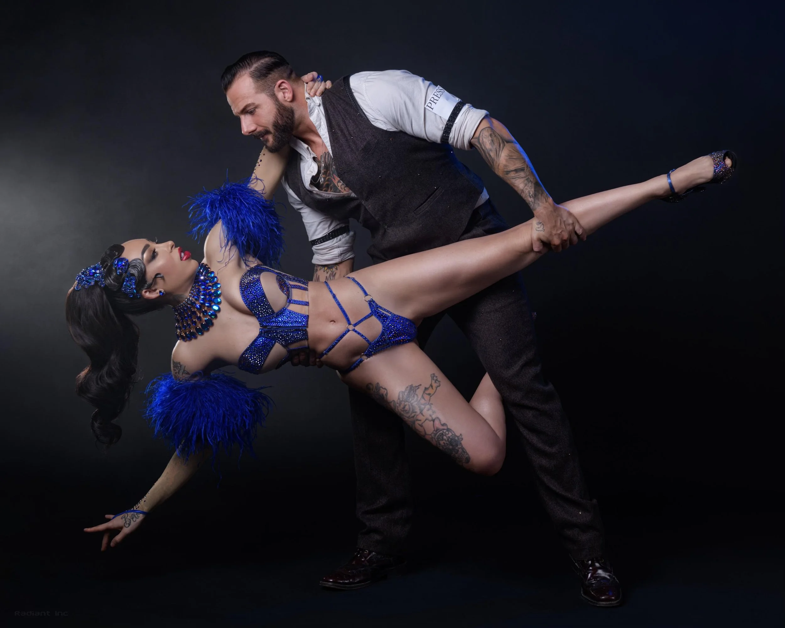 A man and woman dancing, with the man holding the woman by the waist and the woman extending her arm out. The woman is dressed in a blue, rhinestone-studded outfit with feathered accessories, and the man is wearing dark pants and a vest over a white shirt with rolled-up sleeves.