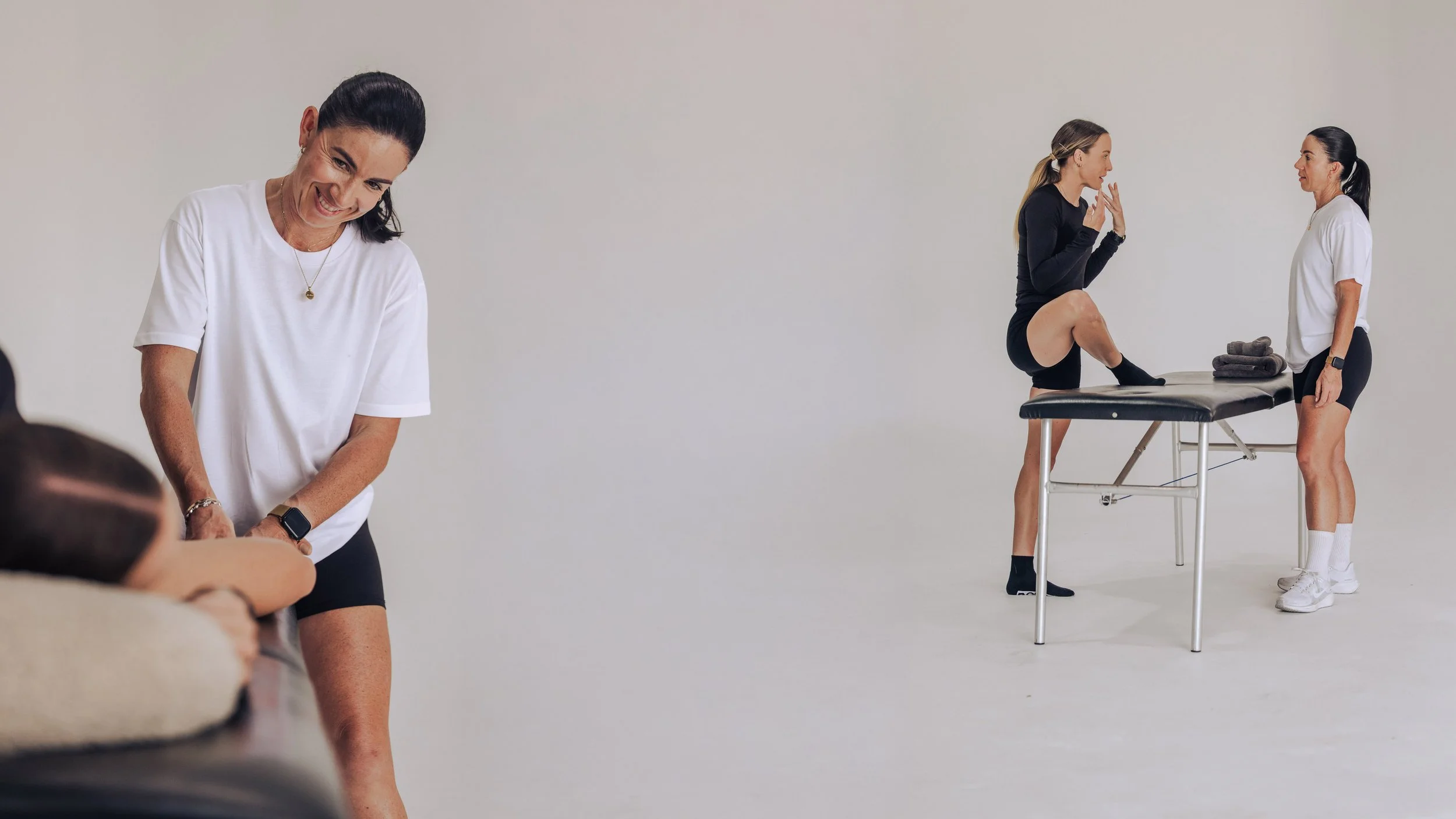 A woman in a white shirt and black shorts smiling while receiving a massage from a therapist, another woman in black athletic wear and socks practicing stretching or yoga on a table with a second therapist standing nearby.