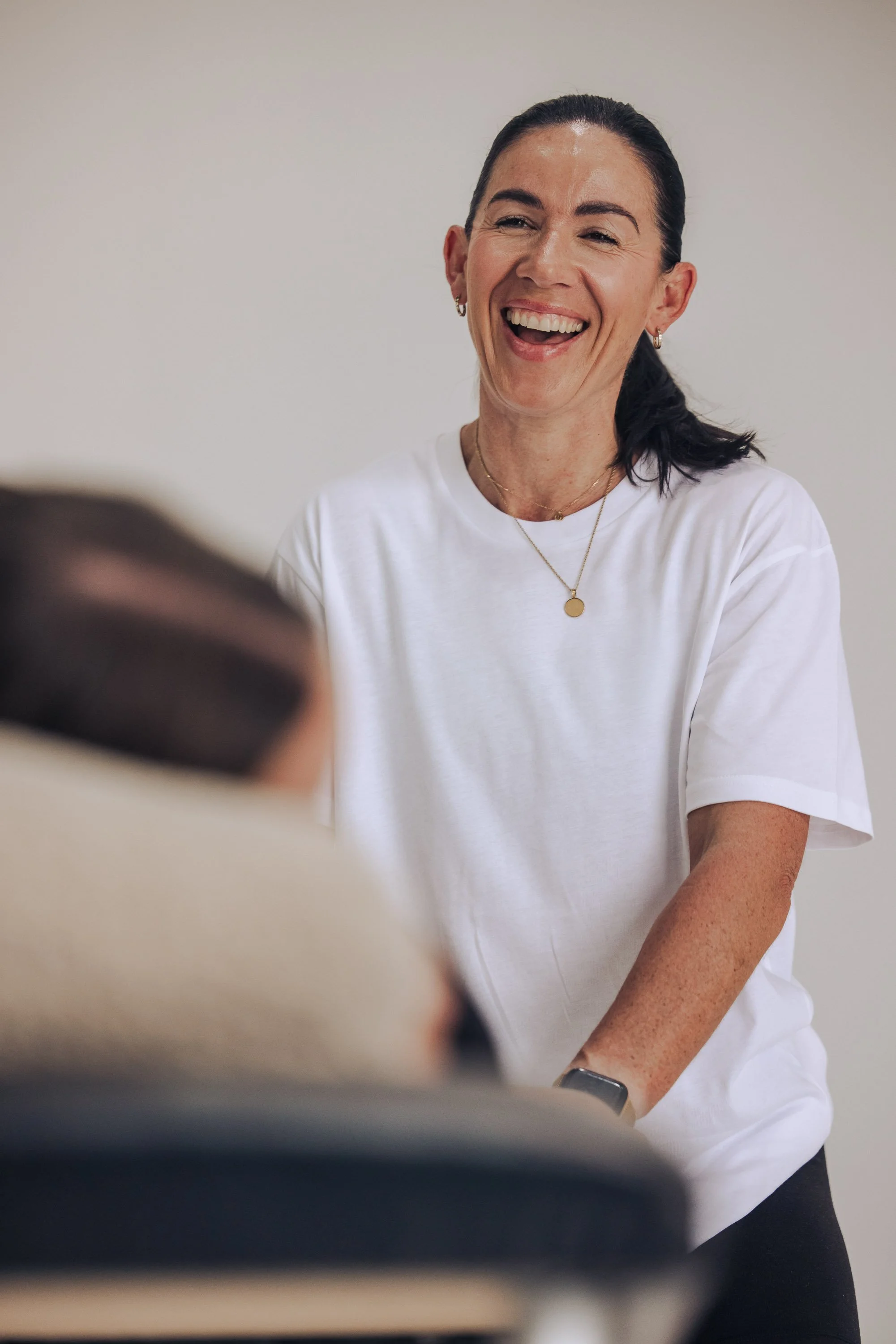 A woman with dark hair pulled back, wearing a white shirt, gold jewelry, and smiling, engaged in conversation.