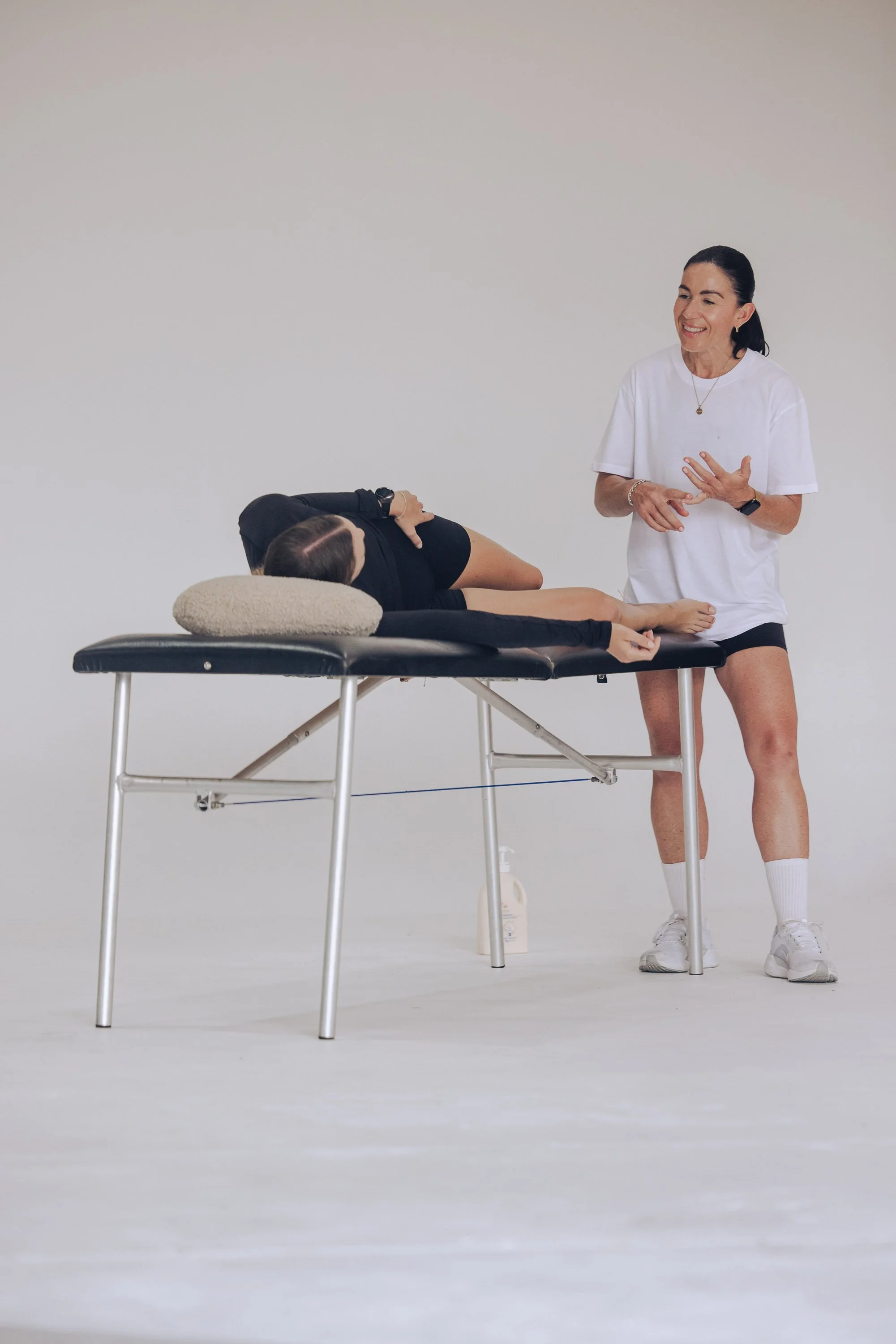 A woman in white athletic clothing instructs a person lying on a massage table, who is resting their head on a pillow. The setting is plain with a white background.