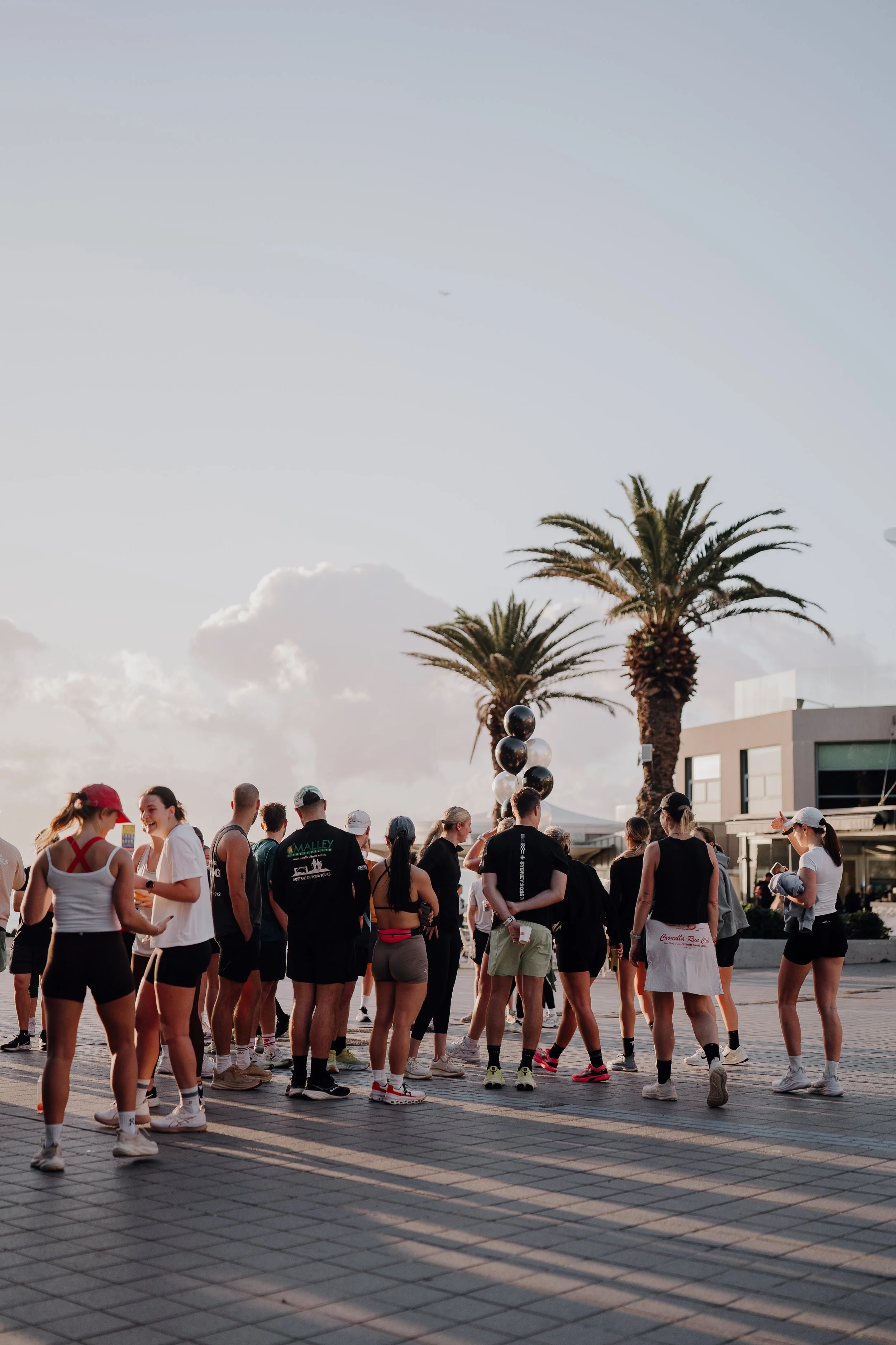 Group of people dressed in athletic wear gathered outdoors, with palm trees and modern buildings in the background during sunset.