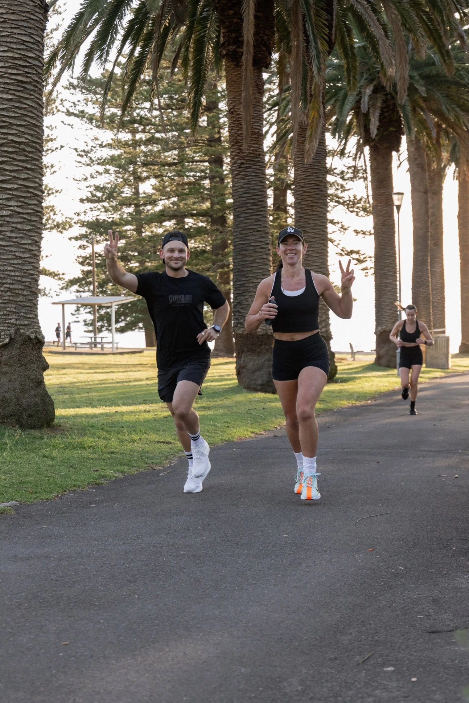 Two people jogging on a park trail with palm trees in the background at sunset, both making peace signs and smiling.