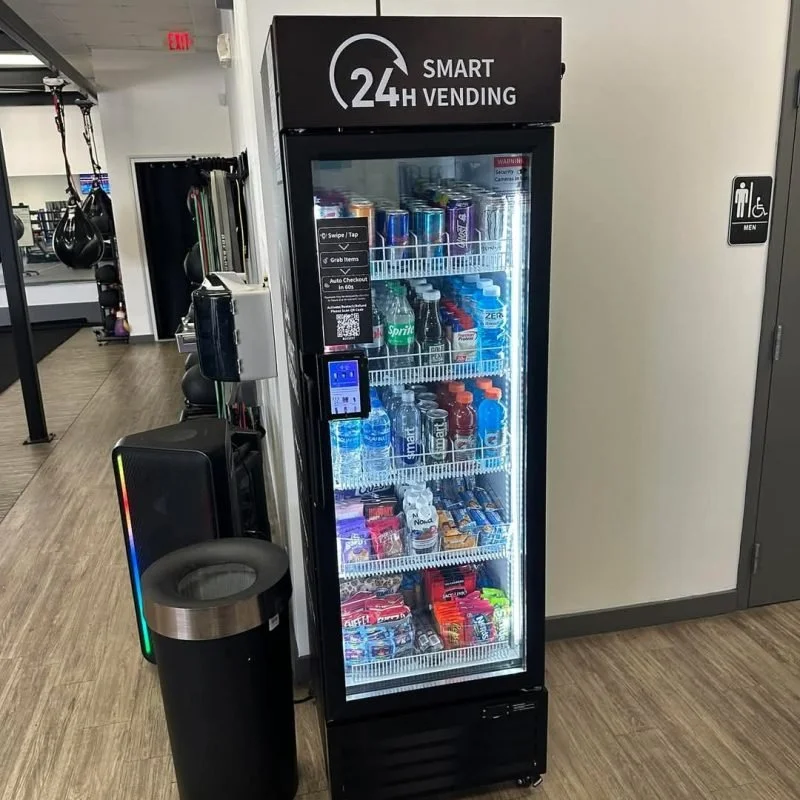 A 24-hour smart vending machine filled with bottled drinks, snacks, and candy, located in a gym facility. It is adjacent to a trash bin and a wall with a men's restroom sign.