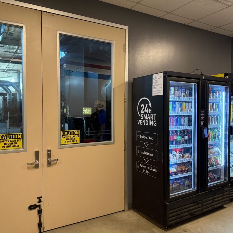 Inside a store, there is a vending machine next to a door with safety glass. The vending machine offers 24-hour smart vending options, including swipe/tap, grab items, and auto checkout in 60 seconds. The door has caution signs indicating safety glasses are required in the area.