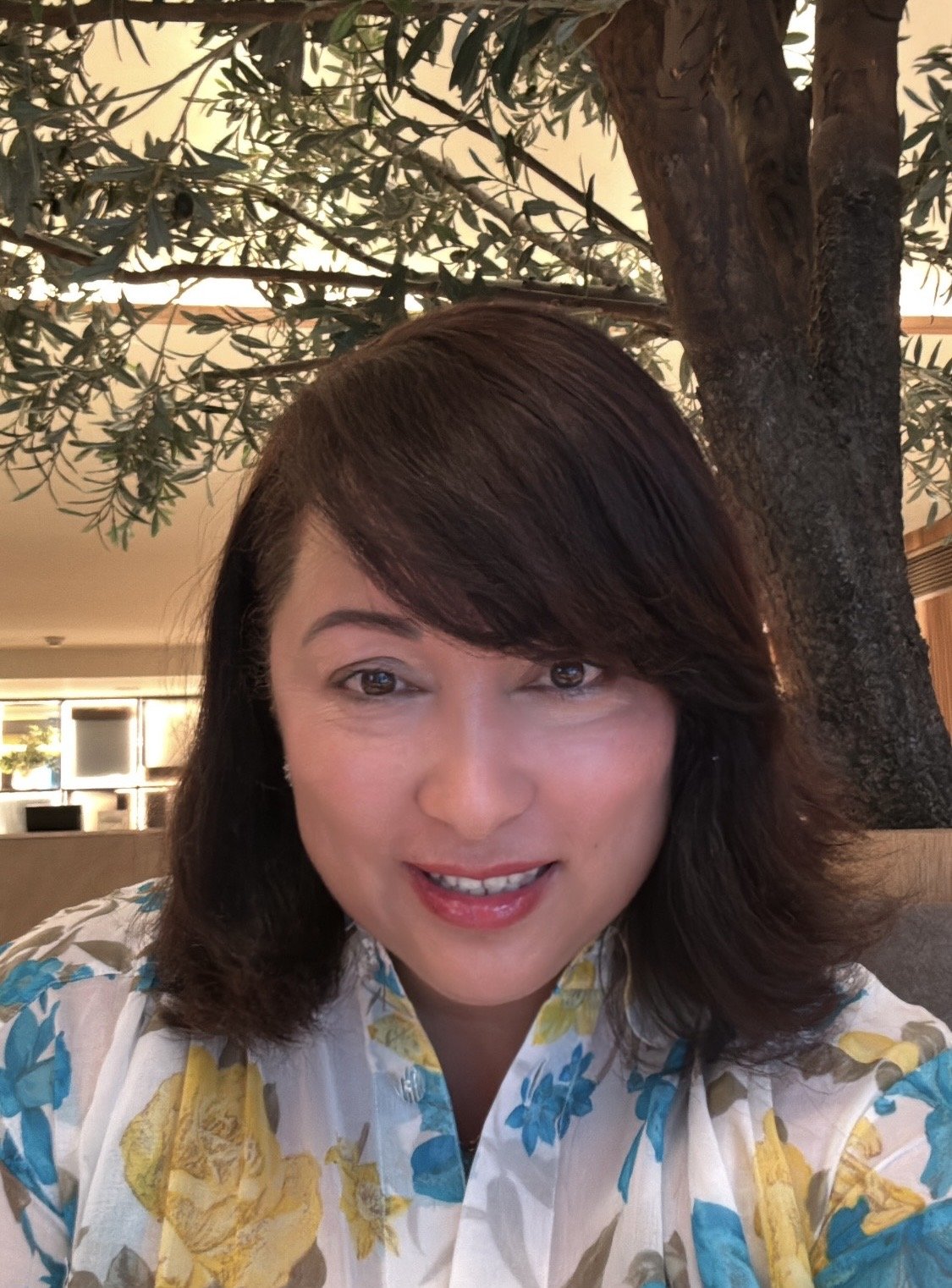 Person smiling in front of a tree indoors, wearing a floral shirt.