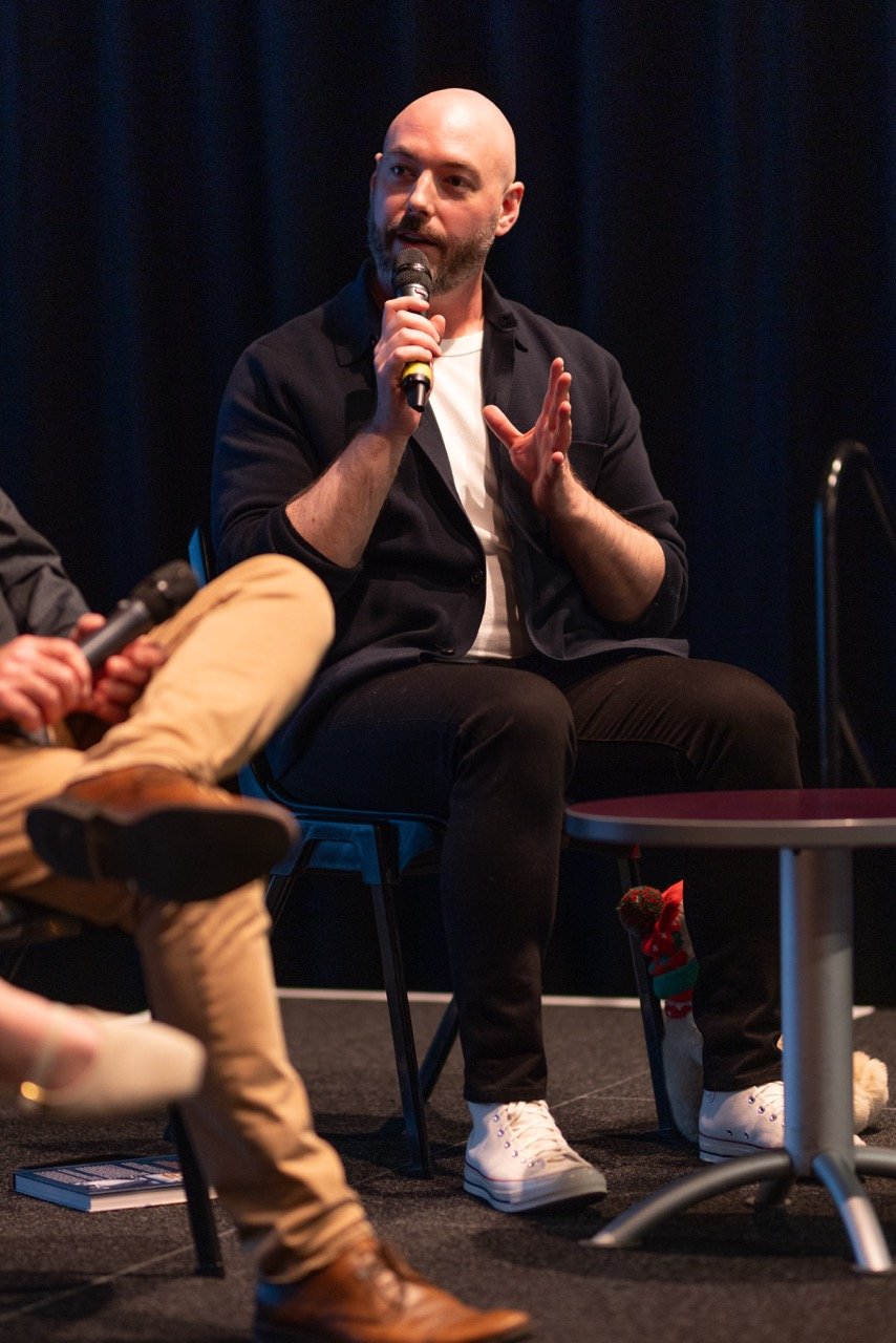 A man with a beard and shaved head speaking into a microphone while sitting on a chair, gesturing with his hand during an event or panel discussion.