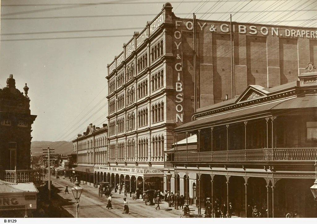 Historic street scene with multi-story brick and wood buildings, including a large building with a sign that says"Foy & Gibson" and "Drapers". People are walking on the sidewalk and horse-drawn carriages are on the street.