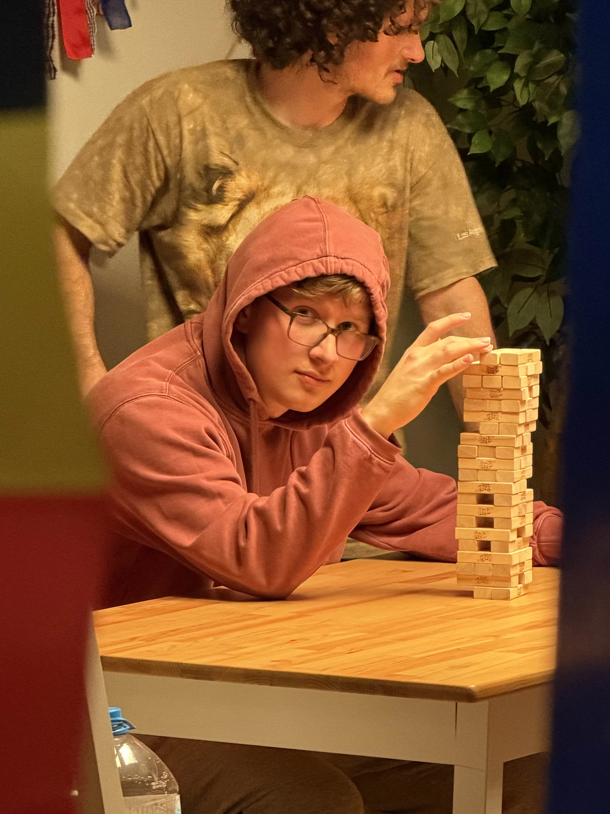 Ethan Page wearing glasses and a pink hoodie is sitting at a table on a short film set, playing Jenga.  The scene is indoors with warm lighting.