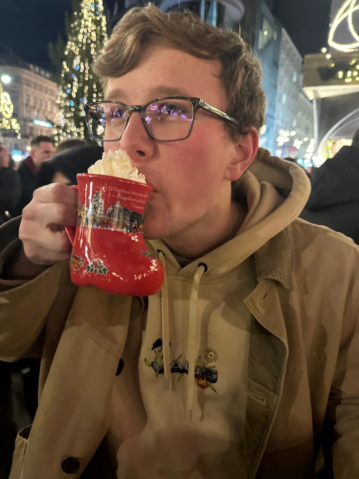 Ethan Page with glasses and light brown hair, wearing a beige jacket and hoodie, is sitting outdoors at night, holding a red Christmas stocking-shaped mug filled with whipped cream, and sipping from it.