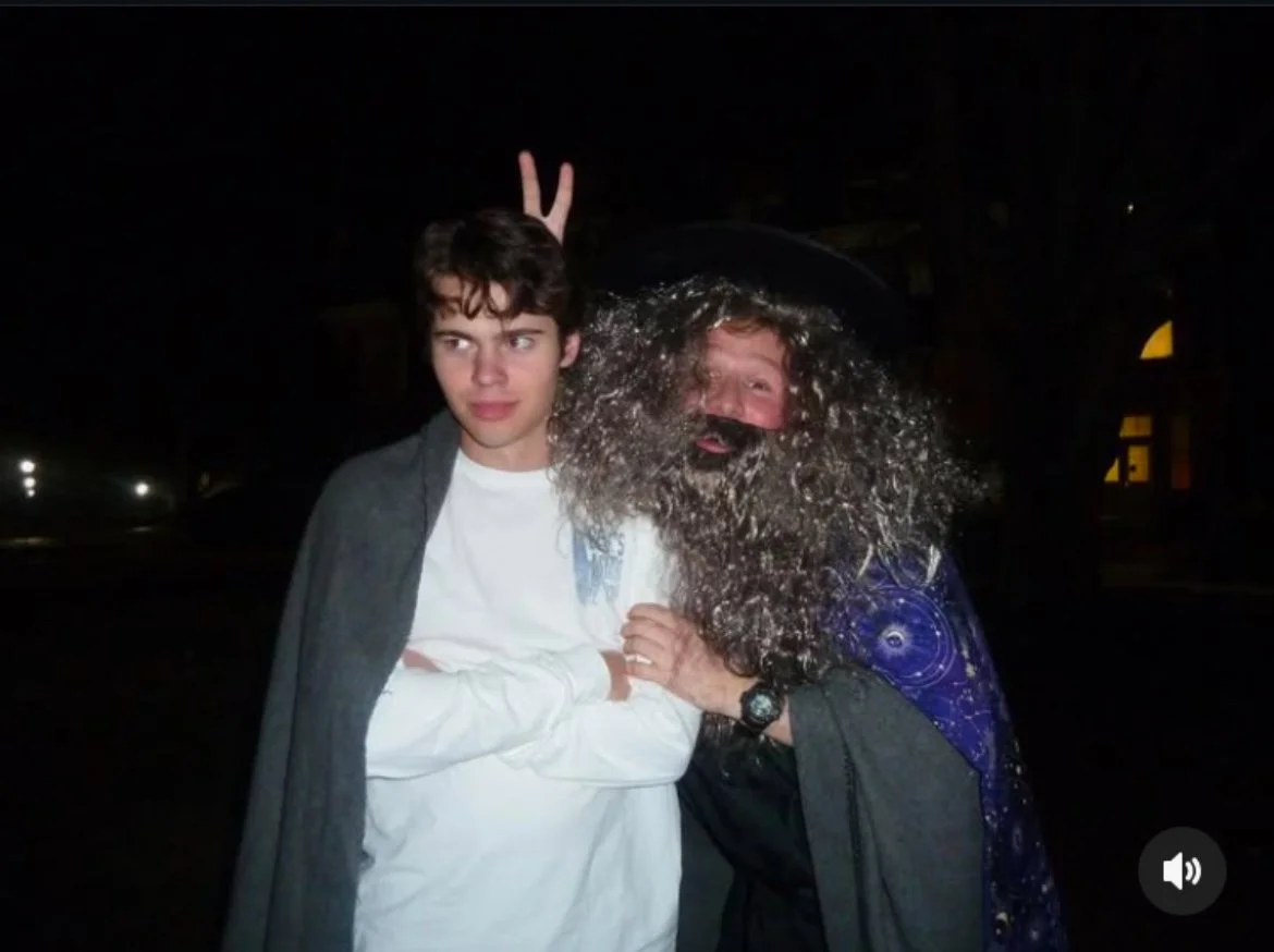 Ethan Page dressed as a wizard with a long, curly beard, a black hat, and a cape with a starry pattern, making a peace sign behind his co-star's head. The co-star has a serious expression, wearing a white shirt and a gray cloak.