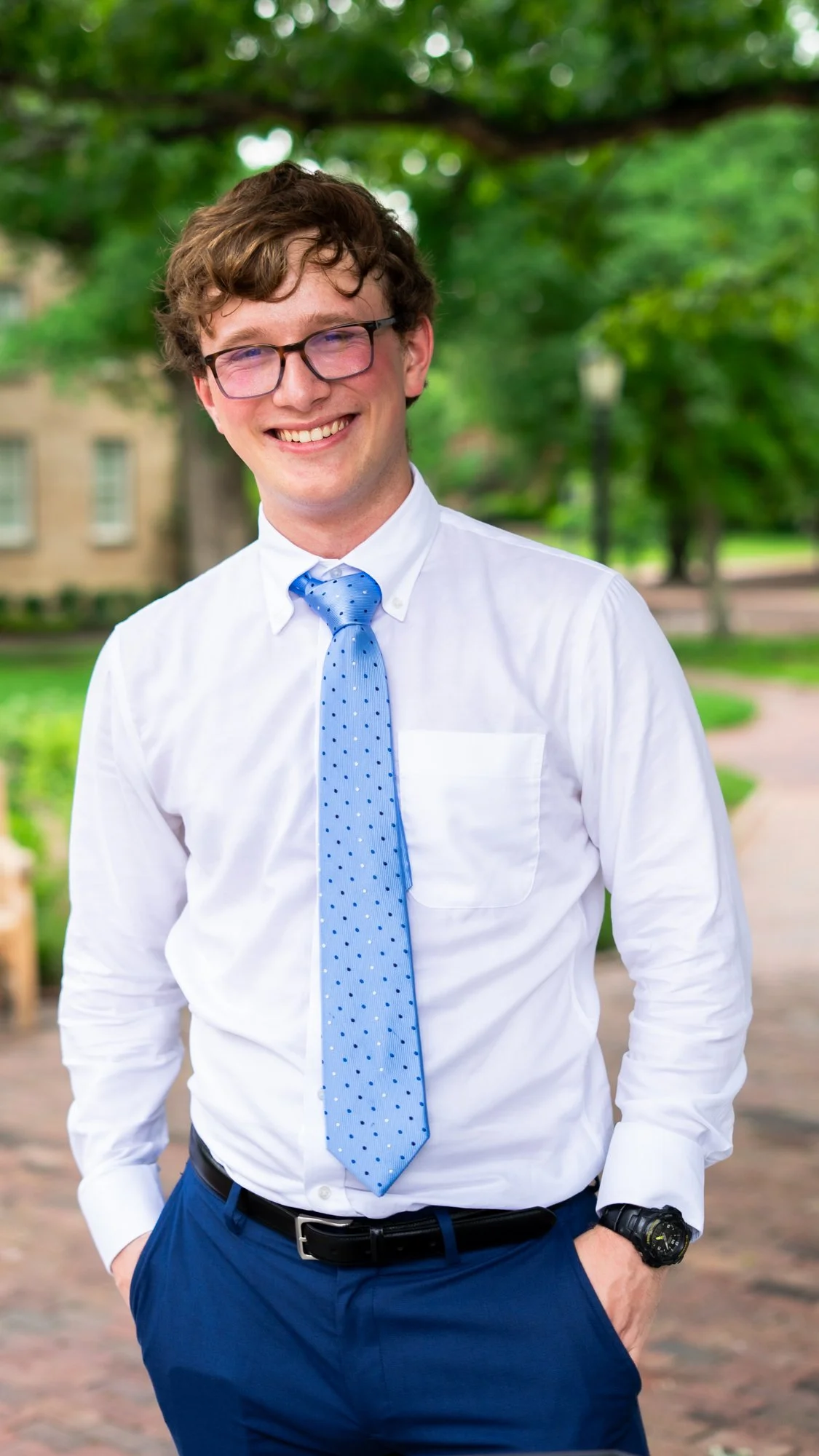 Voice actor Ethan Page with glasses, wearing a white shirt, blue tie with polka dots, and blue pants, smiling outdoors in a park with green trees and a pathway.