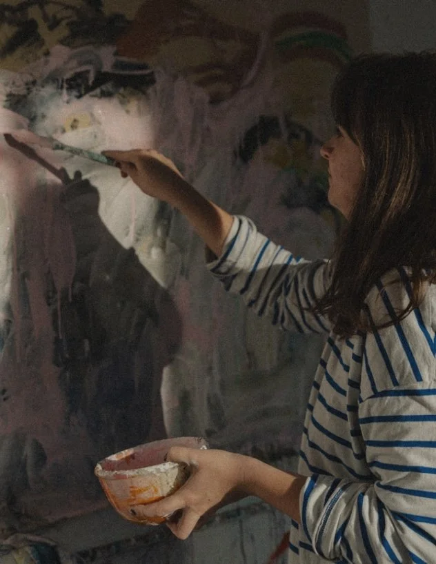 A woman in a striped shirt painting on a large canvas in a dimly lit art studio.