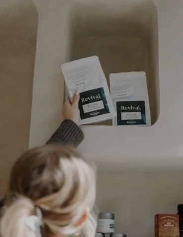 Person with blonde hair in a ponytail reaching into a laundry hamper to grab two packages of Reviva laundry detergent.